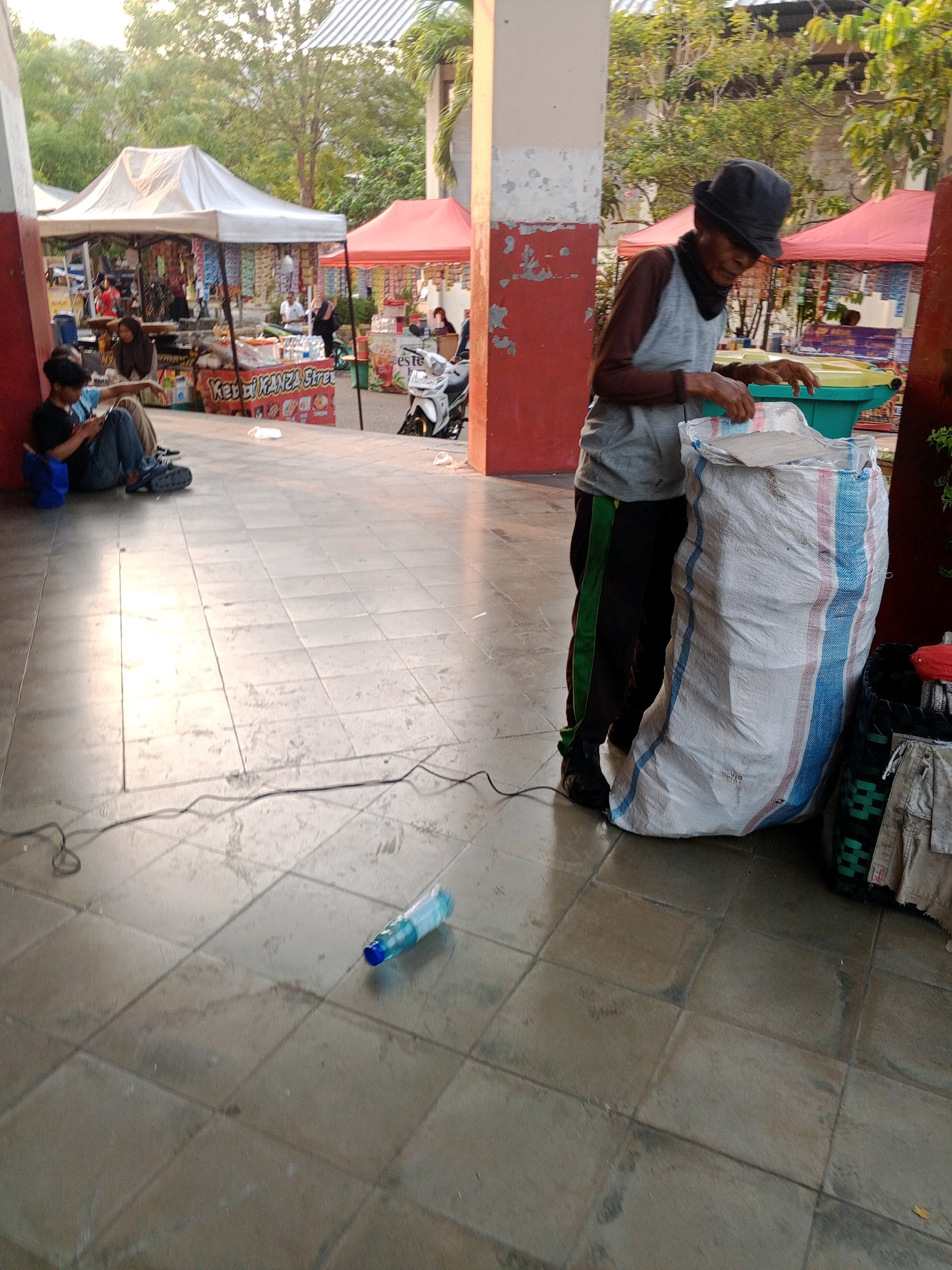 In Wuryorejo, Indonesia, an elderly man wearing a hat and a sleeveless vest over a long-sleeved shirt stands next to a large white bag with blue and red stripes. He is looking down and appears to be sorting through or adding items to the bag, which rests on a tiled floor in front of a structure with a red and white painted pillar. To the left, two individuals are seated on the floor with their backs against the pillar, one of whom is using a mobile phone. In the background, there are several market stalls with white and red canopies, displaying various goods. A white motorcycle is parked near one of the stalls. A plastic water bottle lies on the floor in the foreground, near a black electrical cord. The lighting suggests it is either early morning or late afternoon.