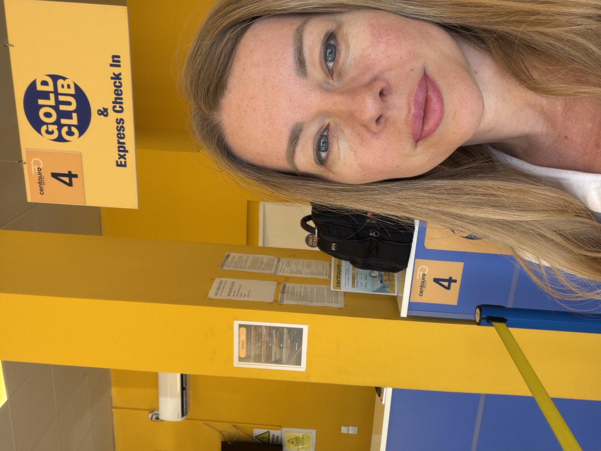 A woman is taking a selfie in front of a brightly colored rental car counter. The counter is predominantly yellow and blue. Above the counter, a yellow sign reads "GOLD CLUB" in large blue letters, with smaller text below that says "& Express Check In". The number "4" is prominently displayed on orange squares on both the sign and on the counter itself, suggesting it is a service bay or queue number. A black backpack is visible on the counter. The woman has blonde hair and is wearing a white shirt. The background suggests an indoor location, possibly an airport or a rental car office in Markopoulo, Greece, given the context provided. The lighting is bright, indicating daytime.