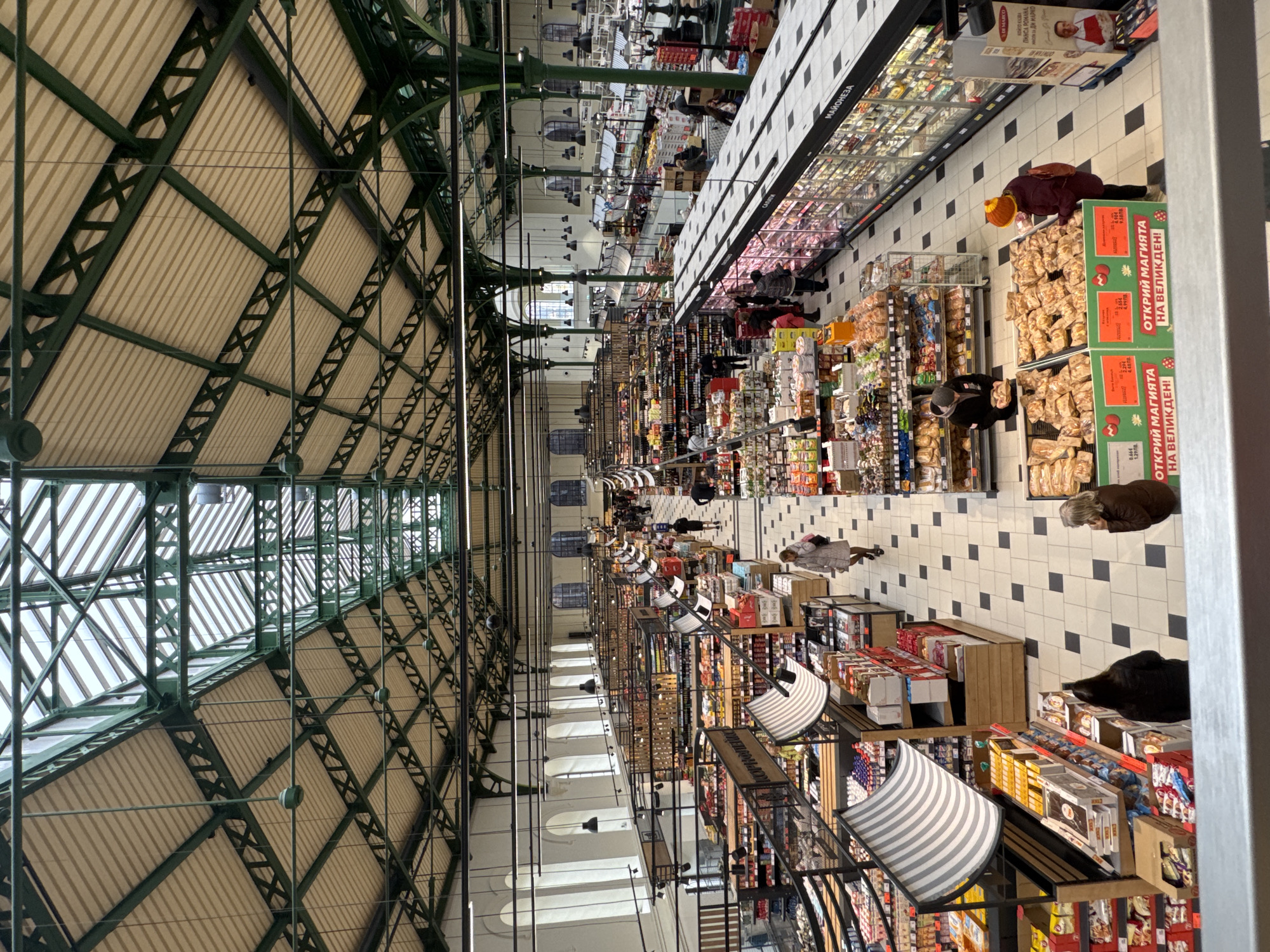 The image shows an interior view of a large grocery store with a high, arched ceiling of steel and corrugated panels. The steel framework is painted a dark green. The floor is tiled in a black and white checkered pattern. The grocery store aisles are long and filled with merchandise. Several people are shopping, some bending over to examine items on the shelves. The text in the image includes prices in Bulgarian currency (ЛВ, or lev), the name "DI MARCO", and Bulgarian phrases, suggesting the location is in Bulgaria. This scene is likely the central market hall in Sofia, Bulgaria.