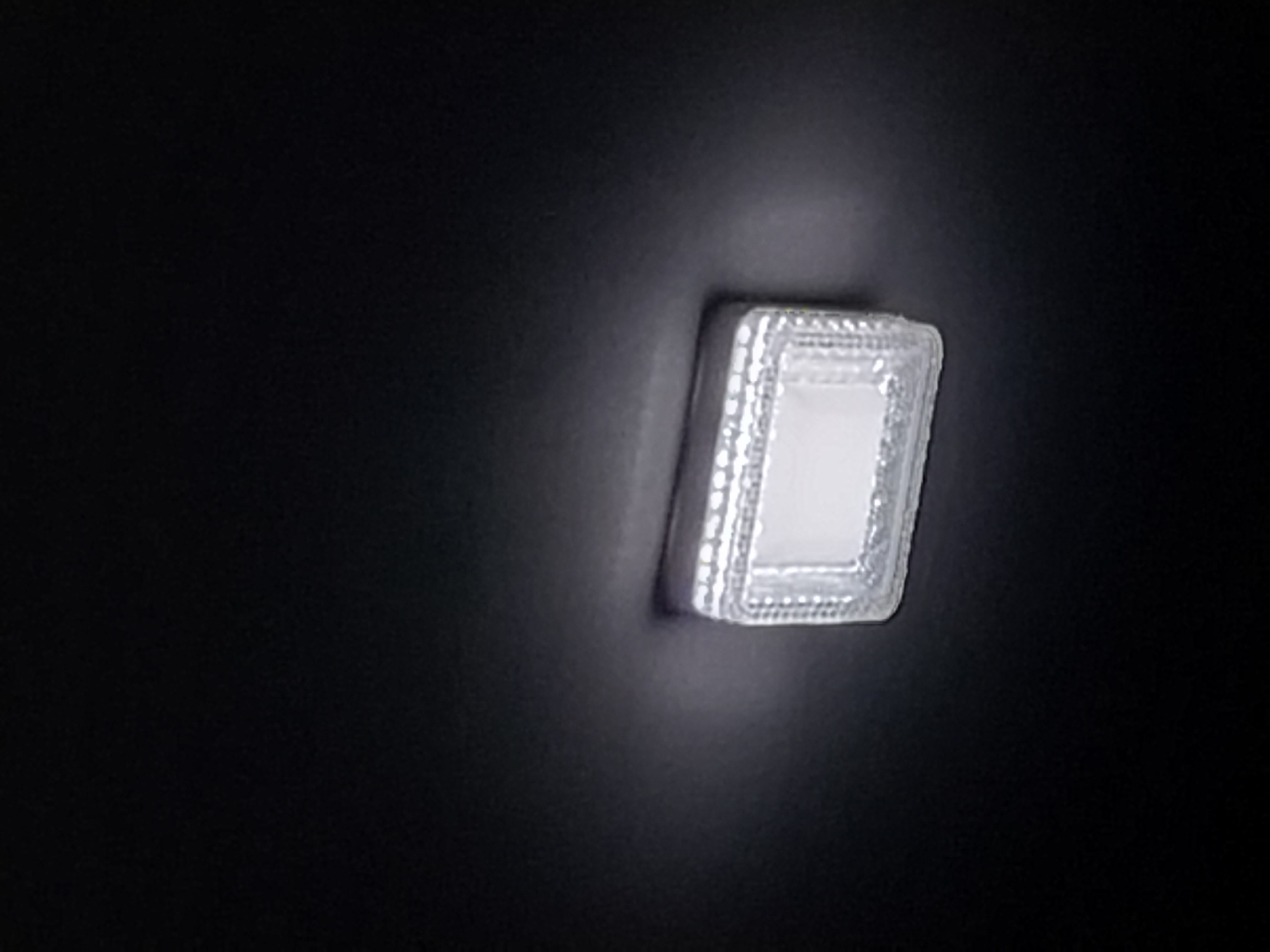 A close-up, low-angle shot shows a small, illuminated rectangular object with a textured, silver-colored frame against a dark, out-of-focus background. The object appears to be a light fixture, possibly an LED panel, embedded in a surface. The frame has a repeating pattern of small, raised dots or spheres. The light emitted from the object is bright white and creates a subtle halo effect on the surrounding dark surface. The scene is devoid of any people, animals, or discernible urban landscape features, making it impossible to determine its location within Dushanbe, Tajikistan. The overall impression is one of a simple, functional light source in a dark environment.