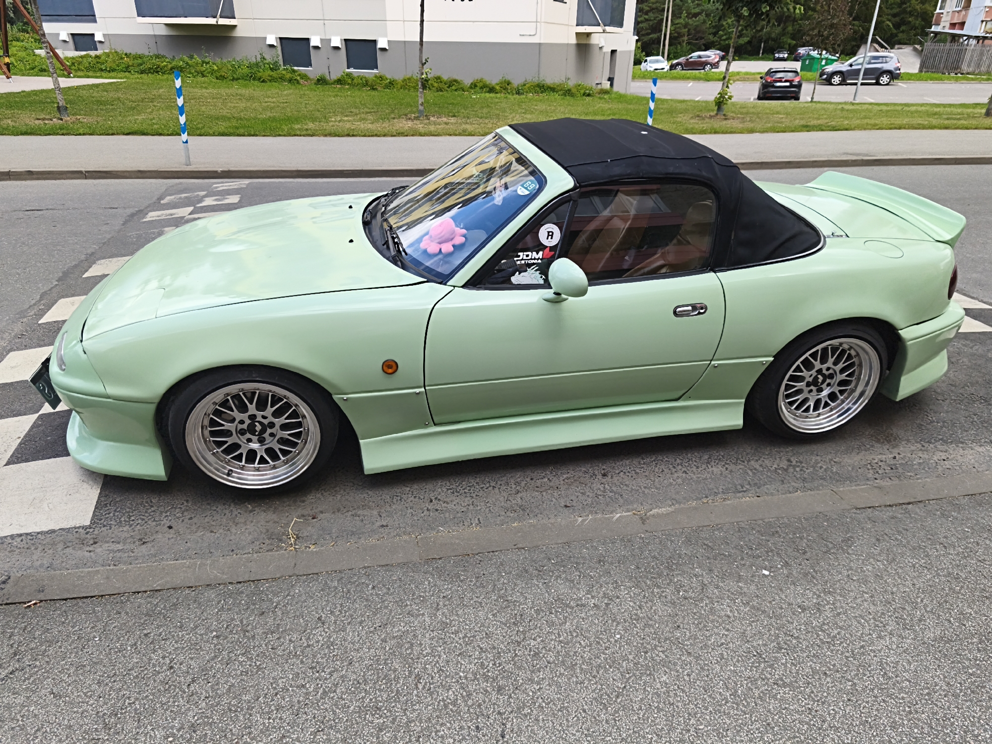 A pale green convertible Mazda Miata with a black soft top is parked on a street in Tallinn, Estonia, near a crosswalk. The car has custom wheels and body modifications, including a spoiler and side skirts. Inside the car, a pink plush toy is visible on the dashboard. The background shows a building with a gray facade, a grassy area, and a parking lot with several other vehicles. The weather appears to be overcast.