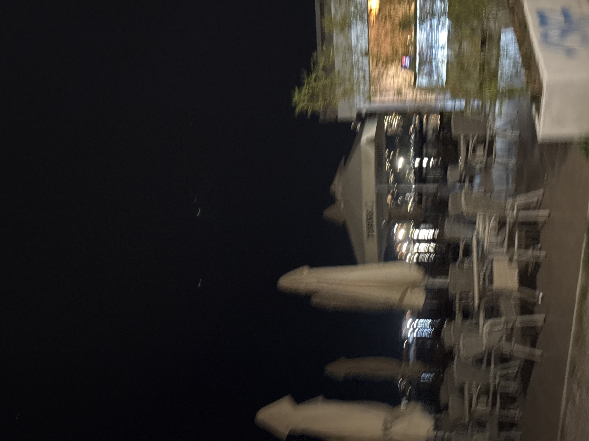 This is a night-time, slightly out-of-focus photograph taken at an outdoor seating area of a restaurant or cafe in Budva, Montenegro. The scene depicts several empty tables and chairs arranged under large, cream-colored umbrellas, suggesting a commercial establishment. In the background, there are blurred lights from buildings and possibly water, indicating a waterfront location or a street with illuminated structures. A white planter with some greenery is visible in the lower right corner, and a portion of a building with a brick-like texture is also present. The overall ambiance is calm and quiet, with the dark night sky serving as the backdrop.