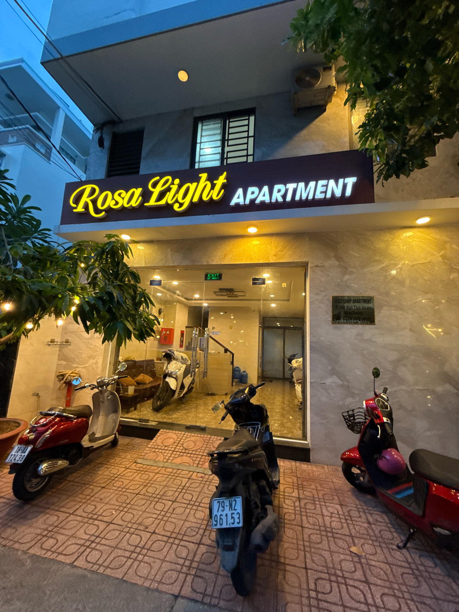 A nighttime exterior shot of the Rosa Light Apartment in Nha Trang, Vietnam, shows the building's entrance and immediate surroundings. The building features a prominent, illuminated sign that reads "Rosa Light APARTMENT" in yellow script and white block letters, respectively. The entrance is a glass-fronted doorway leading into a well-lit interior where several motorcycles are parked. Outside the entrance, three scooters are parked on a patterned tiled sidewalk. The scooter on the left is red and white with license plate "10-AA 124.38". The scooter in the center is dark-colored with license plate "79-N2 961.53". The scooter on the right is red with a black basket and a pink helmet. Trees with string lights are visible to the left of the entrance. A sign for "ROSA LIGHT APARTMENT" with an address and phone number is mounted on the building's wall to the right of the entrance. An air conditioning unit is mounted on the wall above and to the right of the main sign. The sky is dark, indicating it is evening or night.