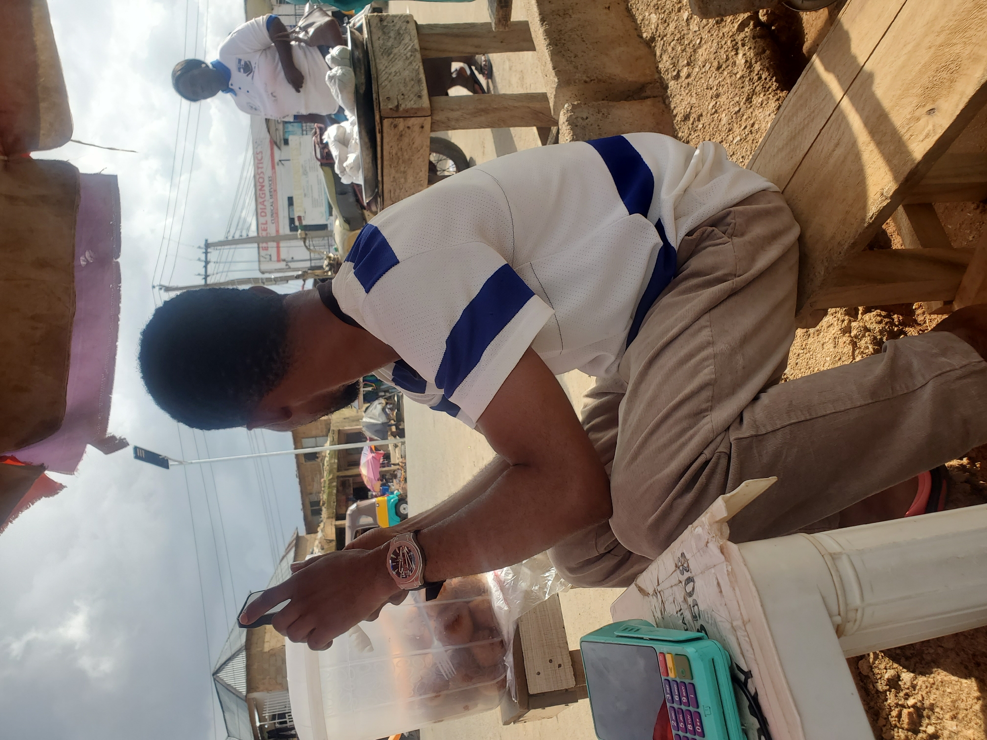 This media file depicts an outdoor scene in Gwong, Nigeria. The primary subject is a young Black man, kneeling and looking intently at his smartphone. He is wearing a white t-shirt with blue stripes and light brown trousers. On his left wrist, he wears a silver wristwatch with a black strap. To his right, on a wooden surface, sits a teal-colored electronic device, likely a point-of-sale terminal, and a clear plastic container filled with food items, possibly fried dough.
In the background, another person is visible, wearing a white shirt with a blue collar and a logo. This individual appears to be standing. The setting suggests a market or roadside stall, with wooden structures and a sandy ground. In the distance, buildings and overhead wires indicate an urban or semi-urban environment. A sign with the text "EXCEL DIAGNOSTICS CLINICAL SERVICES" is also visible. The sky is overcast, suggesting it might be daytime, but the weather is not explicitly clear. The overall mood is one of everyday activity, with the young man engrossed in his phone while at what appears to be his place of business.