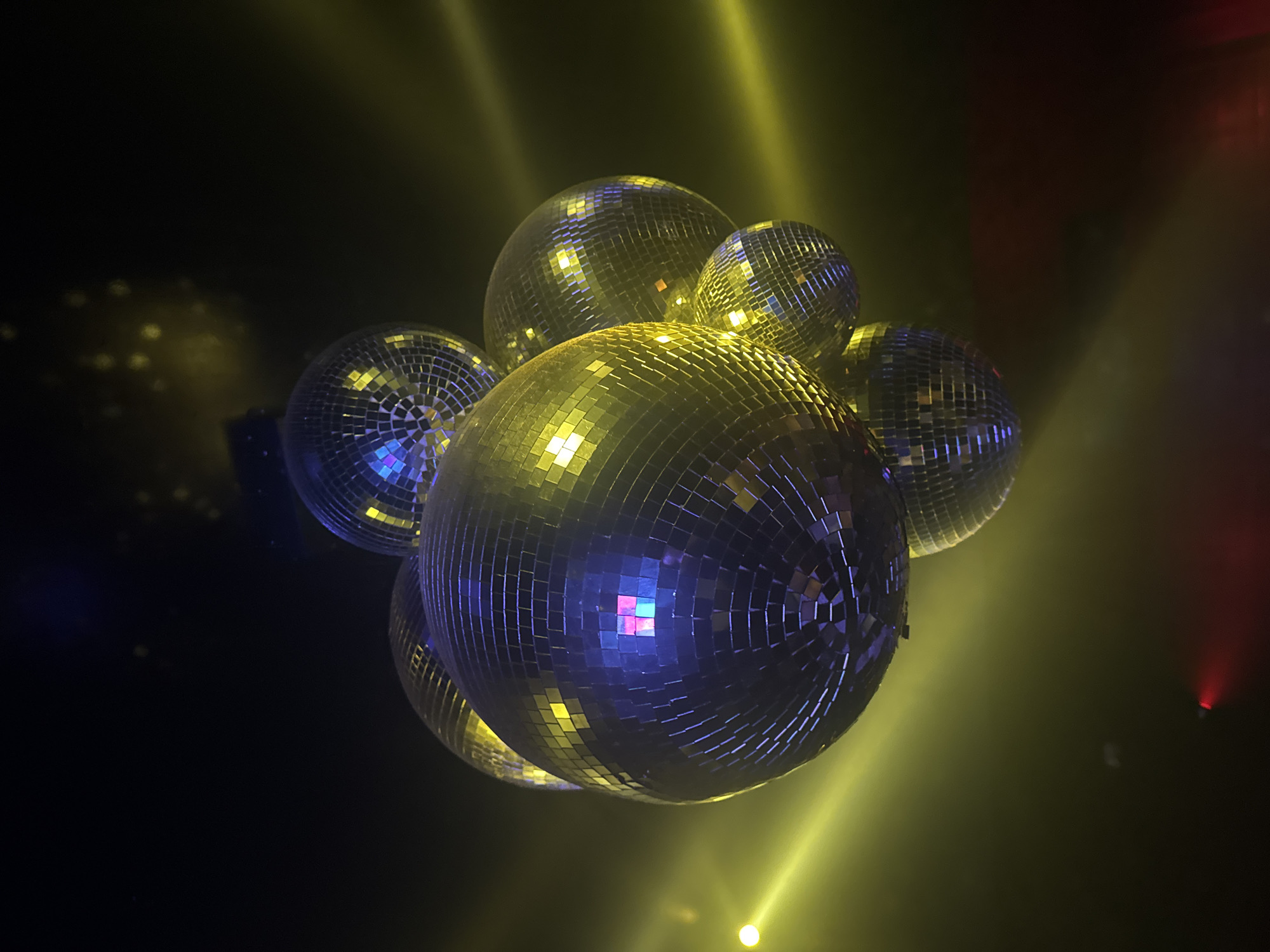 A cluster of seven mirror-covered disco balls hangs suspended against a dark background, illuminated by yellow and purple lights. The disco balls are of varying sizes, with the largest in the center, and are densely packed together. Some of the disco balls reflect the ambient lighting, showing a mosaic of colors. The environment appears to be an indoor space, possibly a nightclub or event venue, with beams of light cutting through a smoky or hazy atmosphere. There is no visible indication of the city being Paris, France.