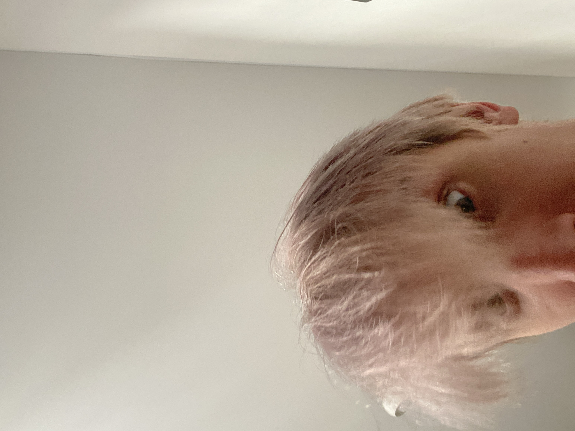 The image is a close-up, low-angle shot of a person's face and head, taken from below. The person has light-colored, possibly dyed, hair that appears somewhat messy. Their left eye is visible and looking towards the camera. The background is a plain, light-colored wall. There is no indication of the city of Sinyavino, Russia, or any other specific location. The overall lighting suggests an indoor environment.