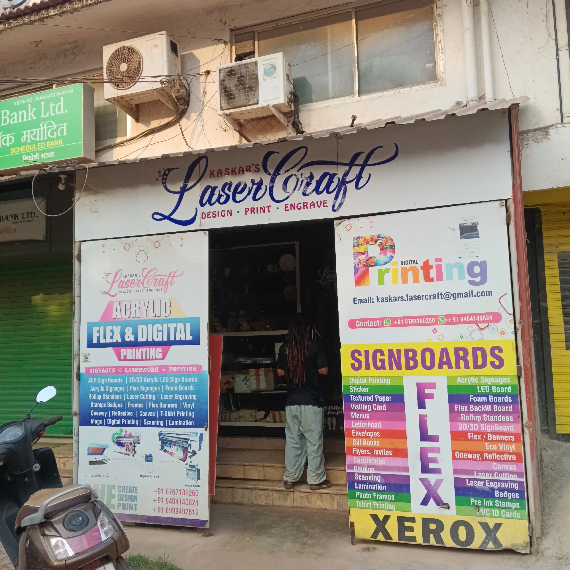 The image depicts the facade of a business called "Kaskar's Laser Craft" in Oxel, India. The shop offers design, print, and engraving services, as indicated by signs displayed on either side of the entrance. These signs advertise acrylic, flex, and digital printing services, along with various signage options like LED boards and foam boards.
The storefront also lists digital printing services, including stickers, textured paper, visiting cards, and more. Contact information, including email and phone numbers, is prominently displayed.
A person with dreadlocks stands in the shop's doorway, viewed from behind. A brown scooter is parked in front of the store. Above the store entrance, there are two air conditioning units and signage for "Bank Ltd.". The building appears to be two stories high.