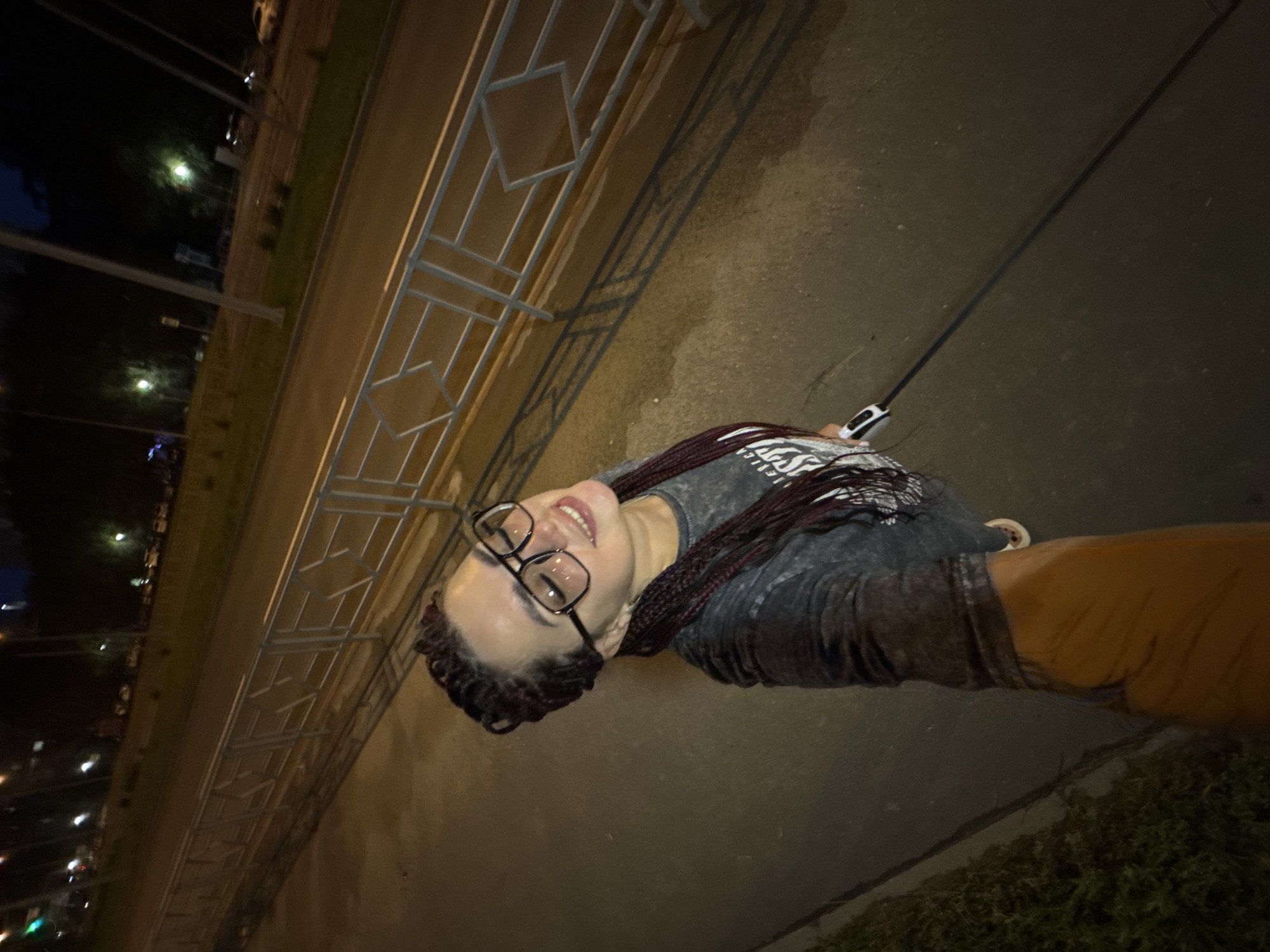 The image shows a young woman with braided hair and glasses, smiling and looking at the camera. She is standing on a sidewalk at night, next to a road with streetlights and some cars. A metal fence runs along the side of the road. The scene is urban, and the lighting suggests it is nighttime. The location is identified as Tolyatti, Russia.