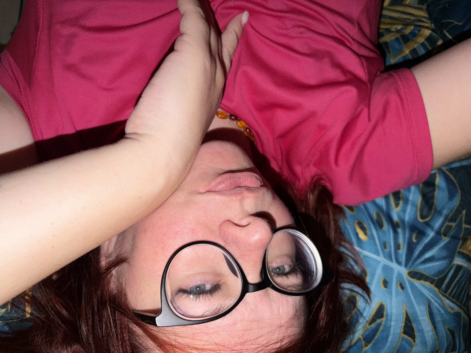 A woman is lying down, presumably on a bed, wearing a reddish-pink shirt and round glasses. She is also wearing a beaded necklace. One of her arms is raised and partially obscuring her face. The background features a blue and gold patterned bedspread. The image does not provide enough information to determine if the picture was taken in the city of Chaikovsky, Russia.