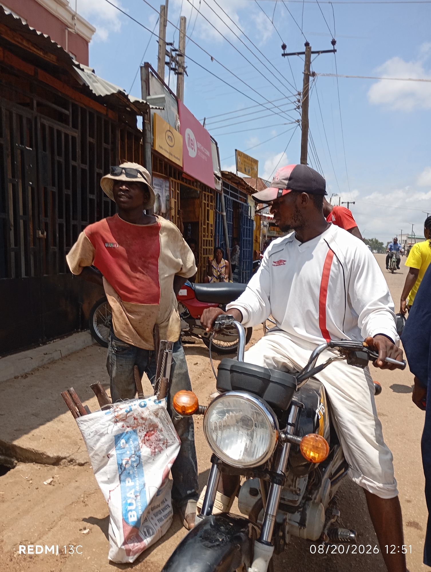 In the city of Shendam, Nigeria, on August 20, 2026, at 12:51 PM, two men are pictured on a street. The man on the left, wearing a wide-brimmed hat and a red and beige shirt, stands with his hands on his hips. In front of him is a white bag filled with wooden poles, and to his right, a red motorcycle is visible. The man on the right, seated on the motorcycle, wears a white long-sleeved shirt with red accents and a cap. He holds the motorcycle's handlebars. The background shows buildings with corrugated metal roofs, a utility pole with wires, and other individuals walking and riding motorcycles in the distance. The ground is dusty and unpaved.