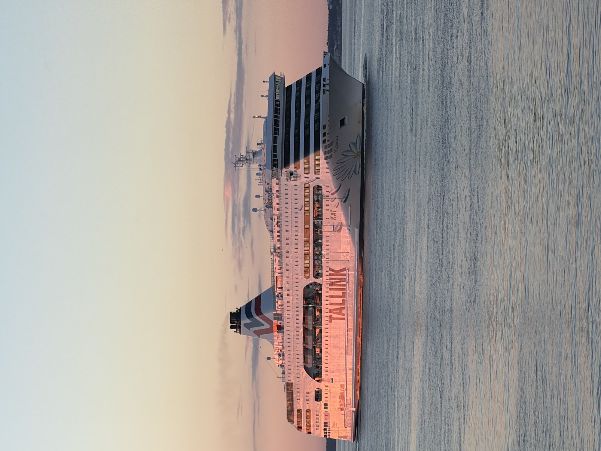 A large white ferry, named "TALLINK VICTORIA I", sails on calm water under a pastel-colored sky at sunset. The ferry is branded with large red lettering spelling out "TALLINK" on its side and a stylized blue flower design near the bow. The ship has multiple decks with many windows. Smoke is visible trailing from one of its funnels. The setting sun casts a warm pink and orange glow on the side of the ferry and reflects on the water's surface. The background shows a faint outline of a coastline with some buildings. This scene is typical of the Tallinn port area in Estonia, where such ferries operate for transport between Tallinn and other Baltic cities. There are no people clearly visible on deck.