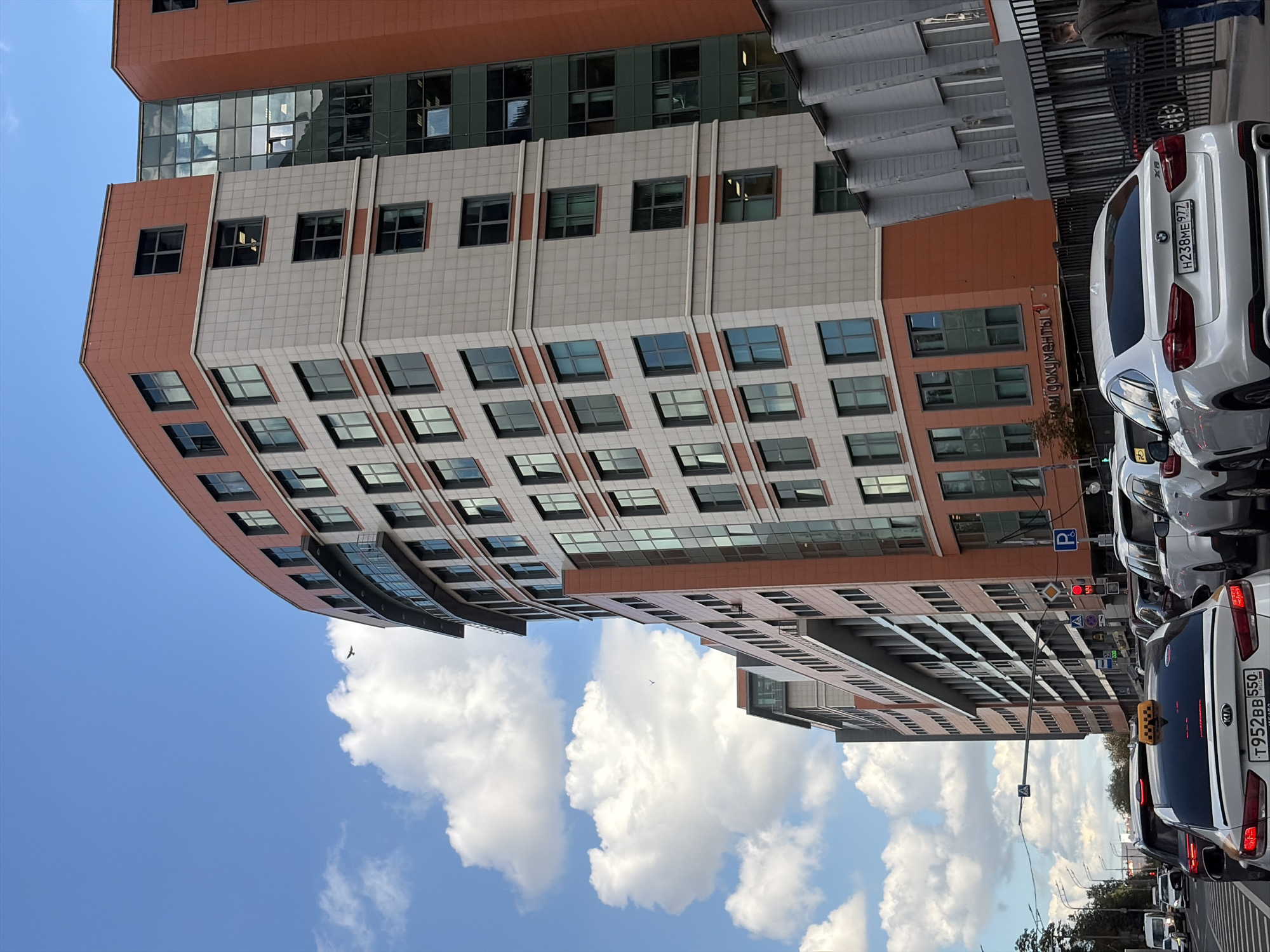 This is a medium-angle, exterior, daytime, eye-level shot of a street scene in Moscow, Russia, captured on a cloudy day. A curved, modern, multi-story building dominates the right side of the frame, featuring an orange-red facade on its upper sections and windows of varying sizes. To the left of this building, another similar structure is visible. In the foreground, a traffic jam is occurring on a multi-lane road. Several cars, including a white Kia sedan with a yellow taxi sign on its roof and a white BMW X6, are stopped in traffic. Pedestrians are not clearly visible, but a person in a jacket and jeans can be seen walking on the sidewalk on the far right. The sky is blue with scattered white clouds, and a bird is visible in flight. The overall environment appears to be an urban setting with modern architecture.
