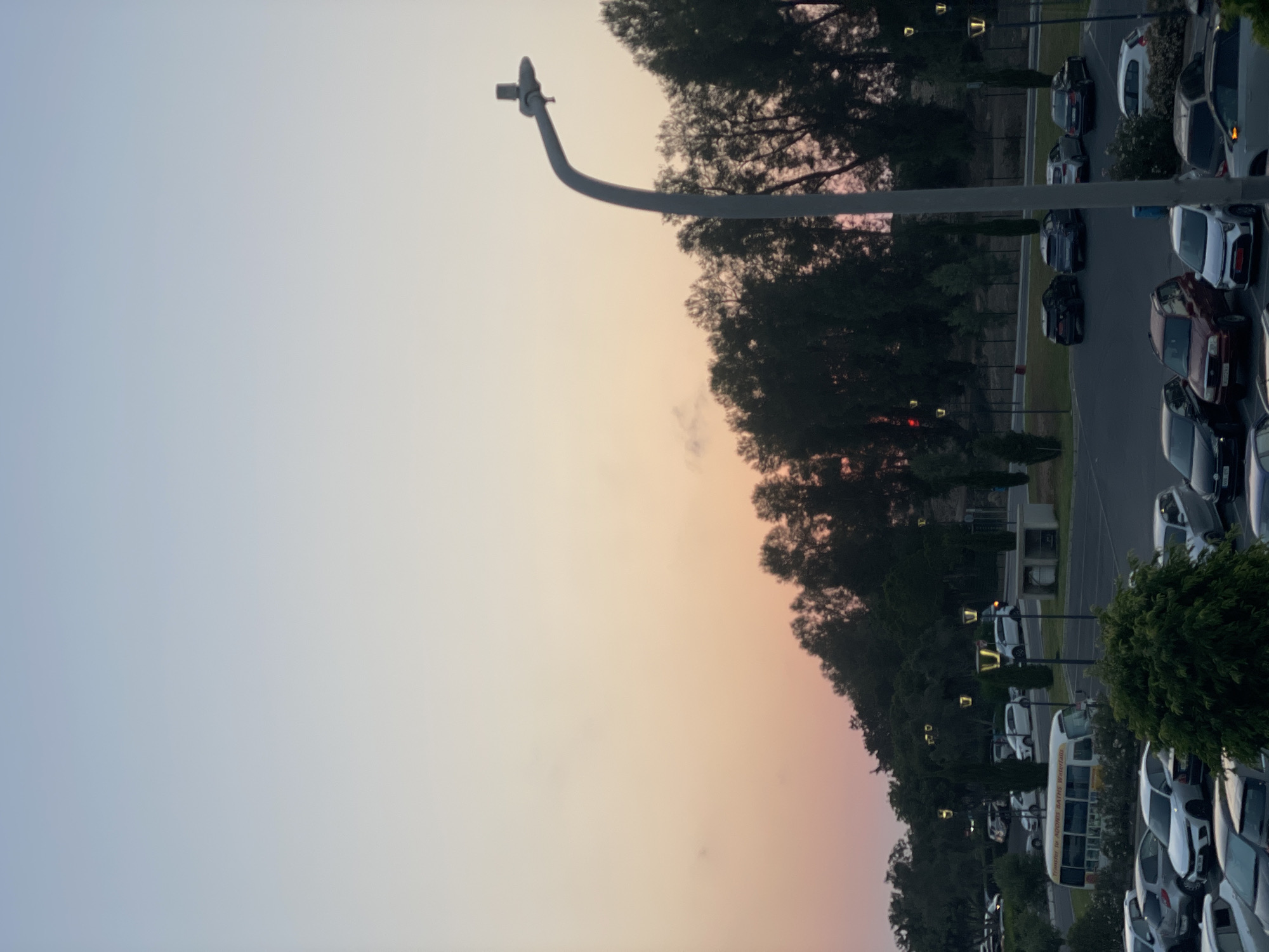 This image captures a parking lot scene during what appears to be either sunrise or sunset, given the soft, gradient sky with hues of pink, orange, and blue. The sky is clear with minimal cloud cover. Dominating the foreground on the left is a tall, grey street lamp with a curved arm. The main focus of the image, however, is the rows of parked cars and a bus that fill the right side of the frame. The vehicles are varied in color and model, suggesting a public or commercial parking area.
Among the vehicles, a white bus with "TRANSIT TO ATRIUM BATES WATERFALLS" written on its side is clearly visible. Several individual cars are parked in designated spots, with some appearing to be sedans and SUVs. The trees lining the parking lot are lush and green, indicating a temperate climate, possibly summer or spring. The ground surface of the parking lot is paved.
The lighting suggests it's either early morning or late evening, casting long shadows and bathing the scene in a warm, soft light. The overall mood is tranquil and calm. There are no people visible in the image. The location is hinted to be in Paphos Municipality, Cyprus, which is known for its historical sites and coastal setting. The presence of a parking lot and a bus suggests a location accessible to visitors or residents, possibly near a tourist attraction or a transport hub.