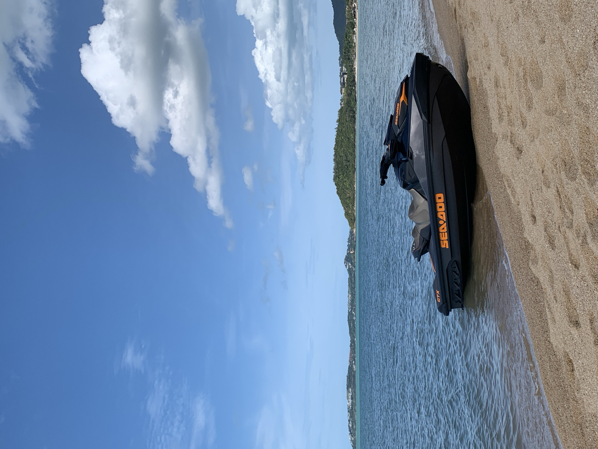 A black Sea-Doo jet ski is parked on a sandy beach, partially in the gentle waves of the ocean. The jet ski is angled towards the right side of the frame. The water is a clear blue-green, and the sand is a light tan color. In the background, across the water, is a coastline with lush green hills dotted with buildings and trees. The sky is a vibrant blue with scattered white clouds. The overall scene appears to be in Baan Bo Phut, Thailand, given the tropical landscape and coastal setting. There are no people visible in the image. The environment is serene and peaceful, suggesting a calm day by the sea.