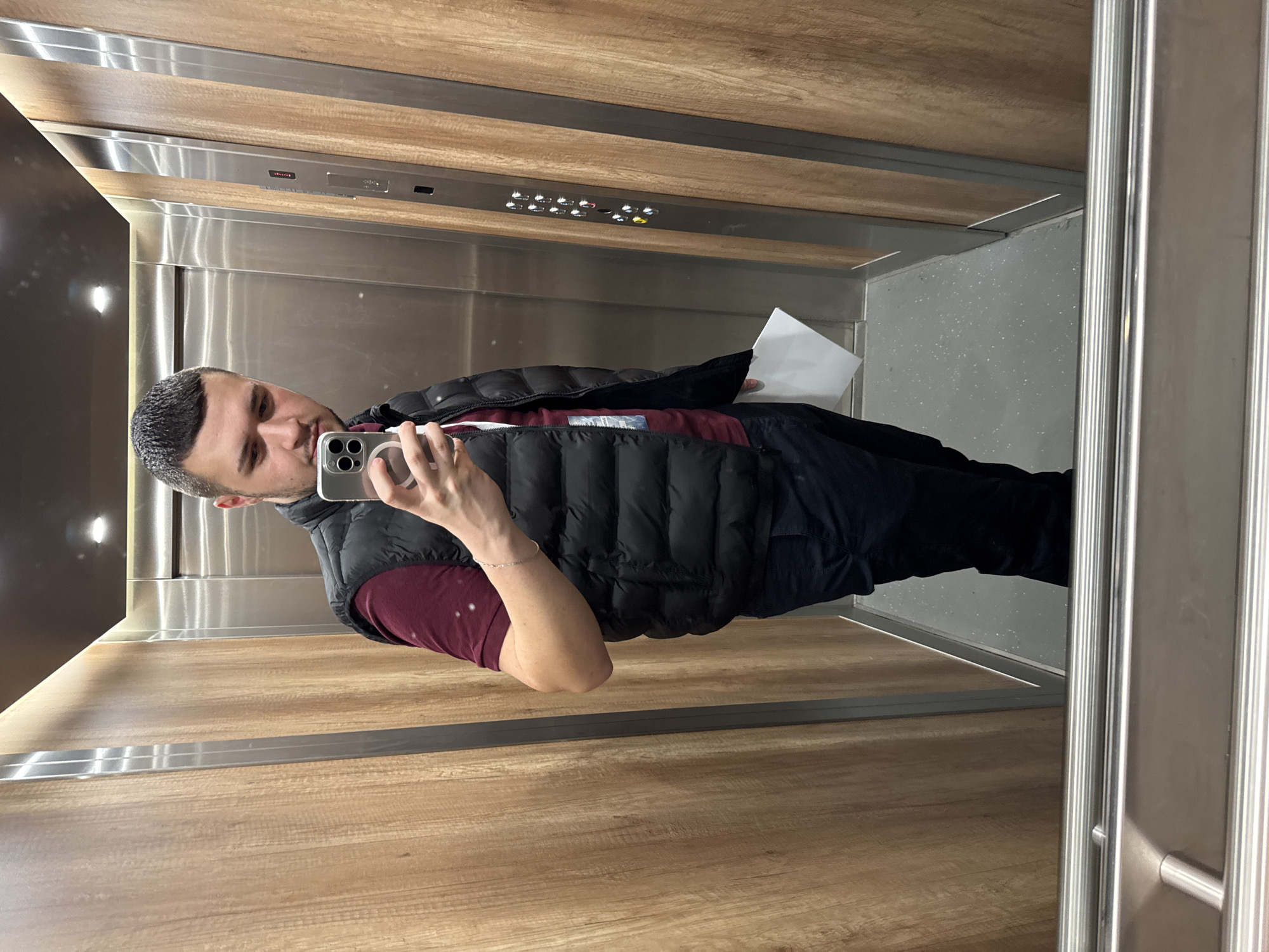 A man is taking a selfie in an elevator with stainless steel walls and a wood-paneled ceiling. The elevator has floor buttons. The man is wearing a maroon shirt, black vest, and dark pants. He is holding a smartphone in his left hand. The elevator door is on the right side of the frame. The photograph could have been taken in Sofia, Bulgaria.