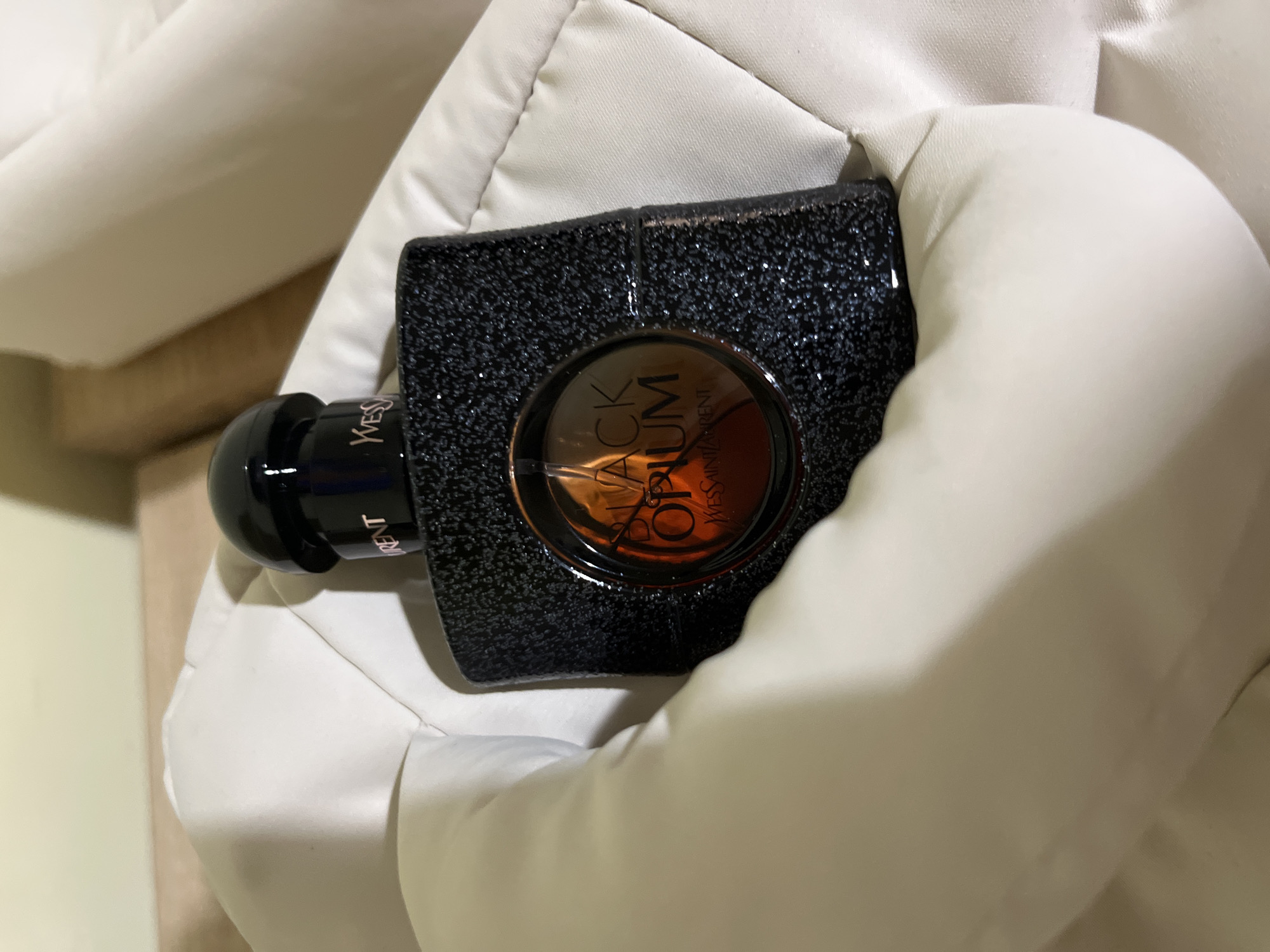 A black, square perfume bottle with a glittery texture is placed on a quilted white surface. The bottle has a black cap and a circular label that reads "BLACK OPIUM" in gold lettering with "YVES SAINT LAURENT" below it. The perfume inside is a dark amber color. The background is a soft, muted yellow.