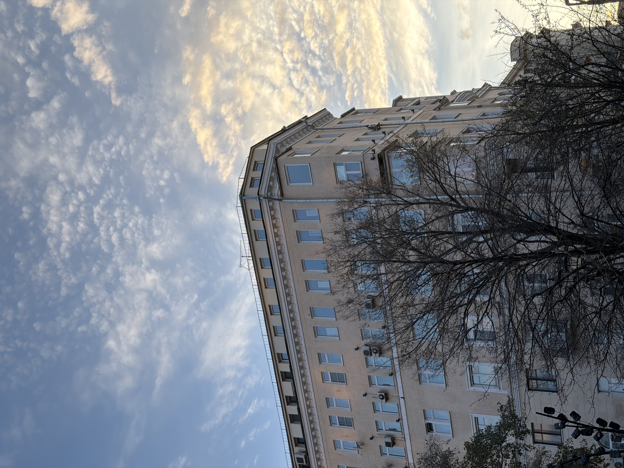 A low-angle shot captures a tall, light-colored building with numerous windows, partly obscured by bare branches of a tree. Several air conditioning units are visible on the building's facade. The sky is filled with textured clouds, displaying a mix of blue and white tones. The location is Moscow, Russia.