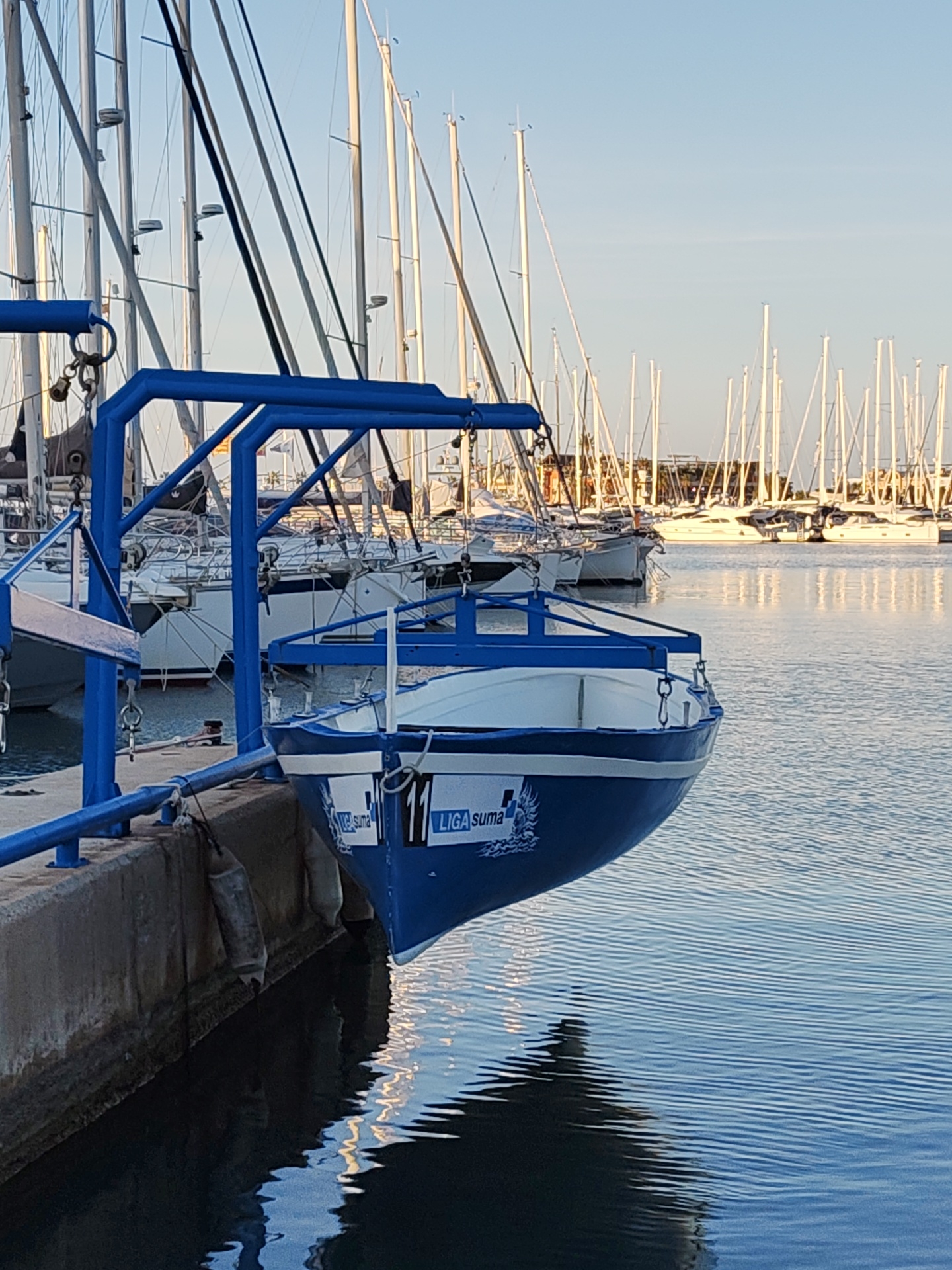 The image depicts a marina scene in Dénia, Spain. In the foreground, a concrete pier edge runs horizontally, with two grey cylindrical fenders hanging from it. A small, blue and white boat, marked with "LIGA suma" and the number "11" on its hull, is suspended just above the water by a prominent, bright blue metal lifting frame or gantry structure. The boat's reflection is visible on the slightly rippled water. In the midground and background, numerous white sailboats and some larger motor yachts are moored. Their vertical masts and rigging form a dense, intricate pattern against a clear, light blue sky. The water surface reflects the boats and sky, displaying subtle light patterns. No individuals are visible in the scene.