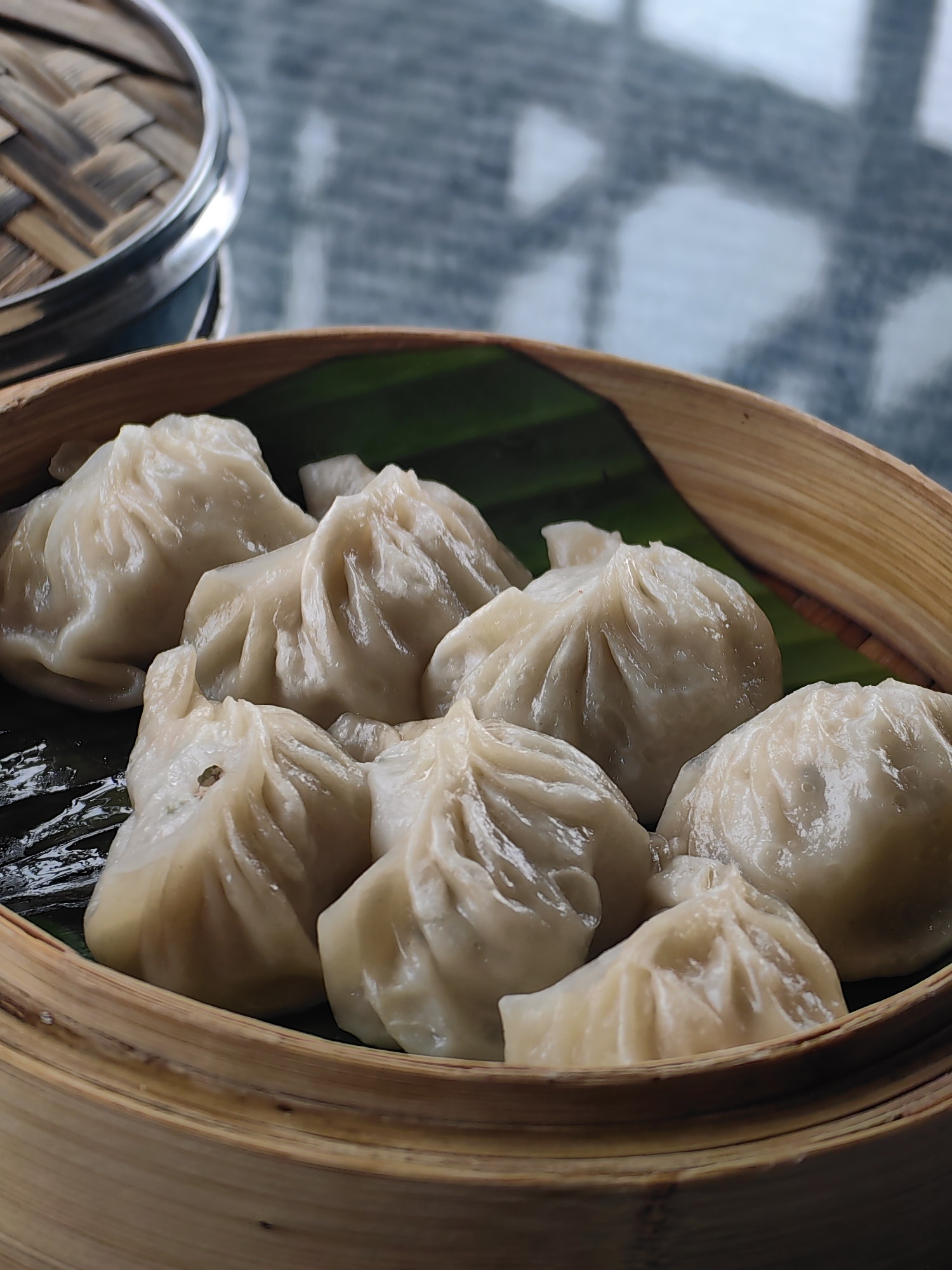 A close-up, eye-level shot showcases a bamboo steamer basket filled with several light-colored dumplings, resting on a dark green leaf. The dumplings have a pleated texture on top. The background is blurred, revealing a surface that appears to be a dark, reflective table, possibly made of granite or a similar stone. In the upper left corner, a portion of another stacked bamboo steamer is visible. The lighting is natural and soft, highlighting the delicate folds of the dumplings. There are no people or animals visible in the image. The scene is static and implies a prepared meal ready to be served. The setting is likely a restaurant or a home dining area in Delhi, India.