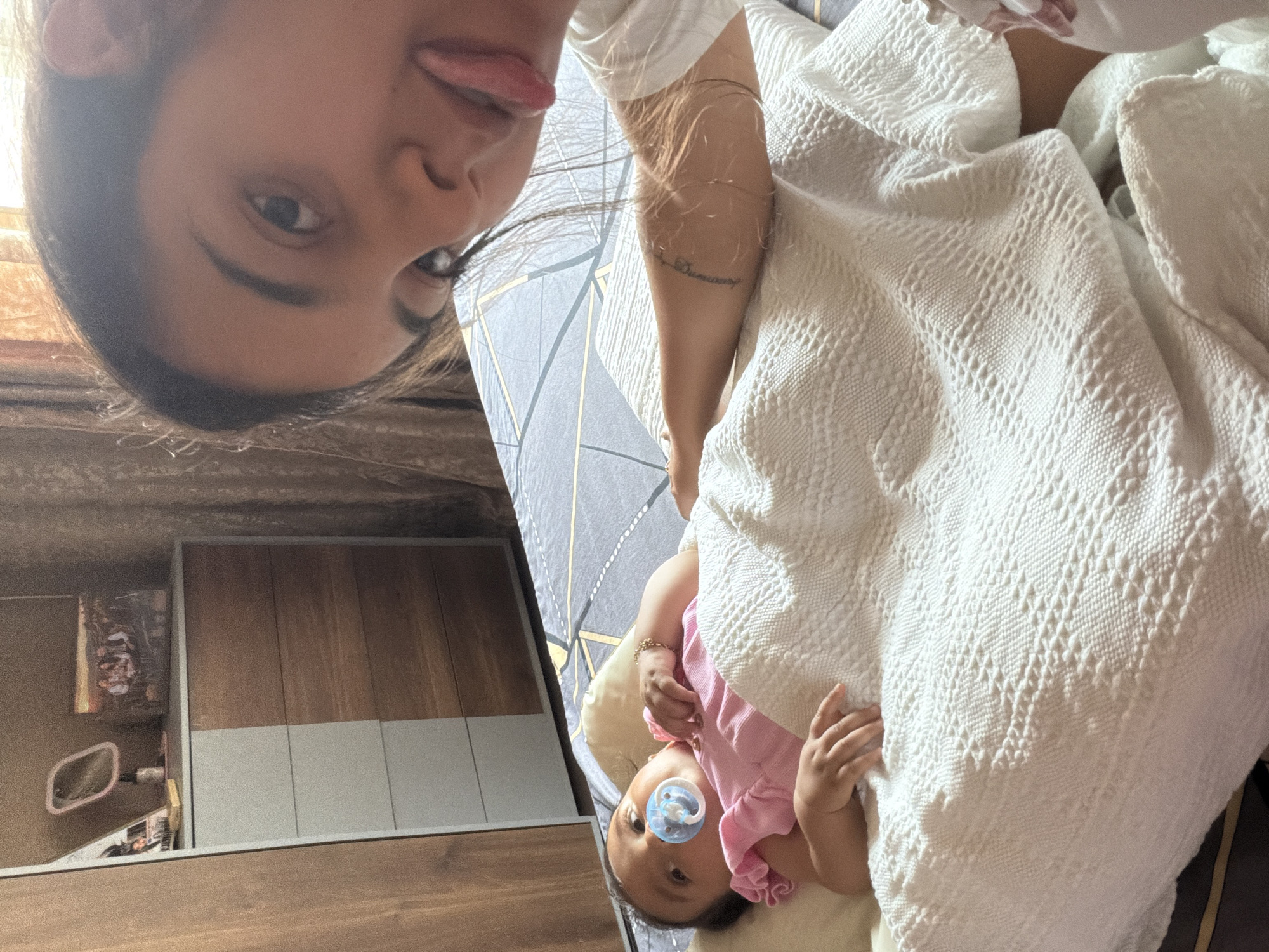 The image shows a close-up selfie with a baby lying on a bed in the background. The person taking the selfie, a young woman, is in the foreground, with her face close to the camera and her tongue sticking out. She is wearing a white t-shirt. In the background, a baby is lying on its back on a bed, wearing a pink outfit and a blue pacifier. The bed has a patterned sheet and is covered with a white textured blanket. Behind the bed, a dresser with a mirror and decorative items is visible, along with a curtain. The setting appears to be a bedroom. The city of Razhevo Konare, Bulgaria, is mentioned as the location, but there are no specific visual cues in the image to confirm this.