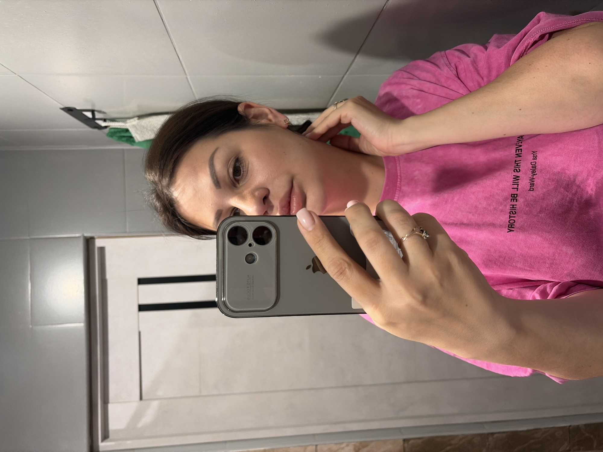 A woman with brown hair is taking a mirror selfie in what appears to be a bathroom. She is wearing a pink t-shirt with text on the back. Her right hand is holding her phone, and her left hand is touching her neck. She is wearing earrings and rings. The bathroom has grey tiled walls and a white door with black vertical stripes. A green towel and a white bag are hanging on a hook on the wall. The image does not contain any information that allows for the determination of the location in the city of Sochi, Russia.