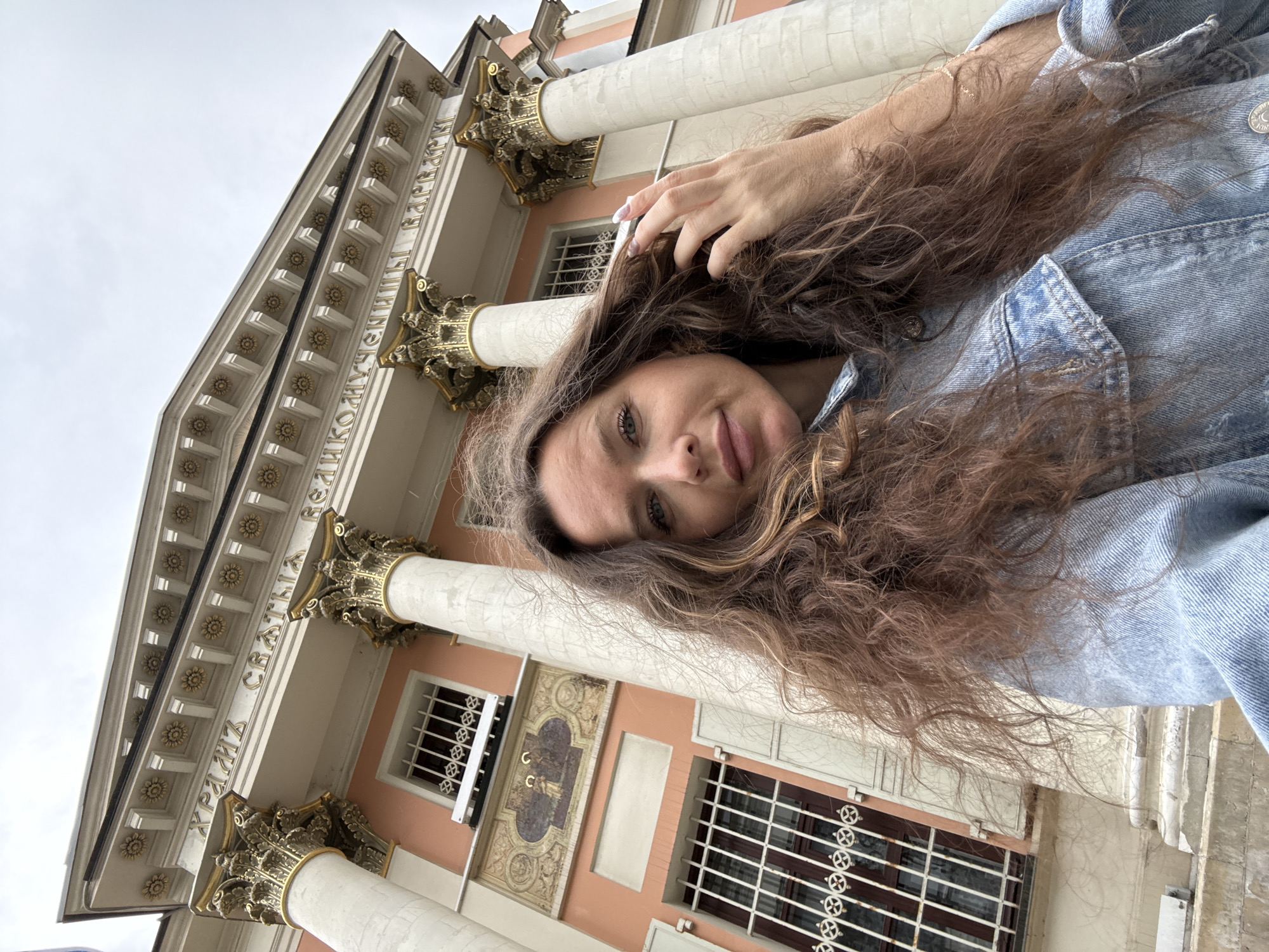 A woman with long, curly brown hair and light blue eyes is taking a selfie in front of a building with classical architecture. She is wearing a light blue denim jacket. The building has a peach-colored facade, white columns with ornate gold capitals, and a pediment with inscribed text in Cyrillic. The text appears to read "ХРАМЪ СВЯТЫЯ ВЕЛИКОМУЧЕНИЦЫ ВАРВАРЫ" which translates to "Church of the Great Martyr Barbara". This church is located in Moscow, Russia. The sky is overcast.
