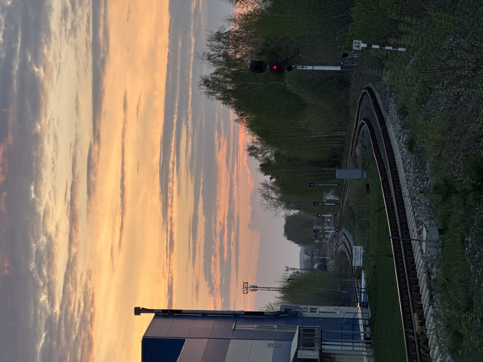 The image depicts a railway track curving into the distance, with a row of railway signal lights visible. A building is in the foreground, with a sunset sky above. The setting appears to be near an industrial or urban area, given the presence of the building and railway infrastructure.