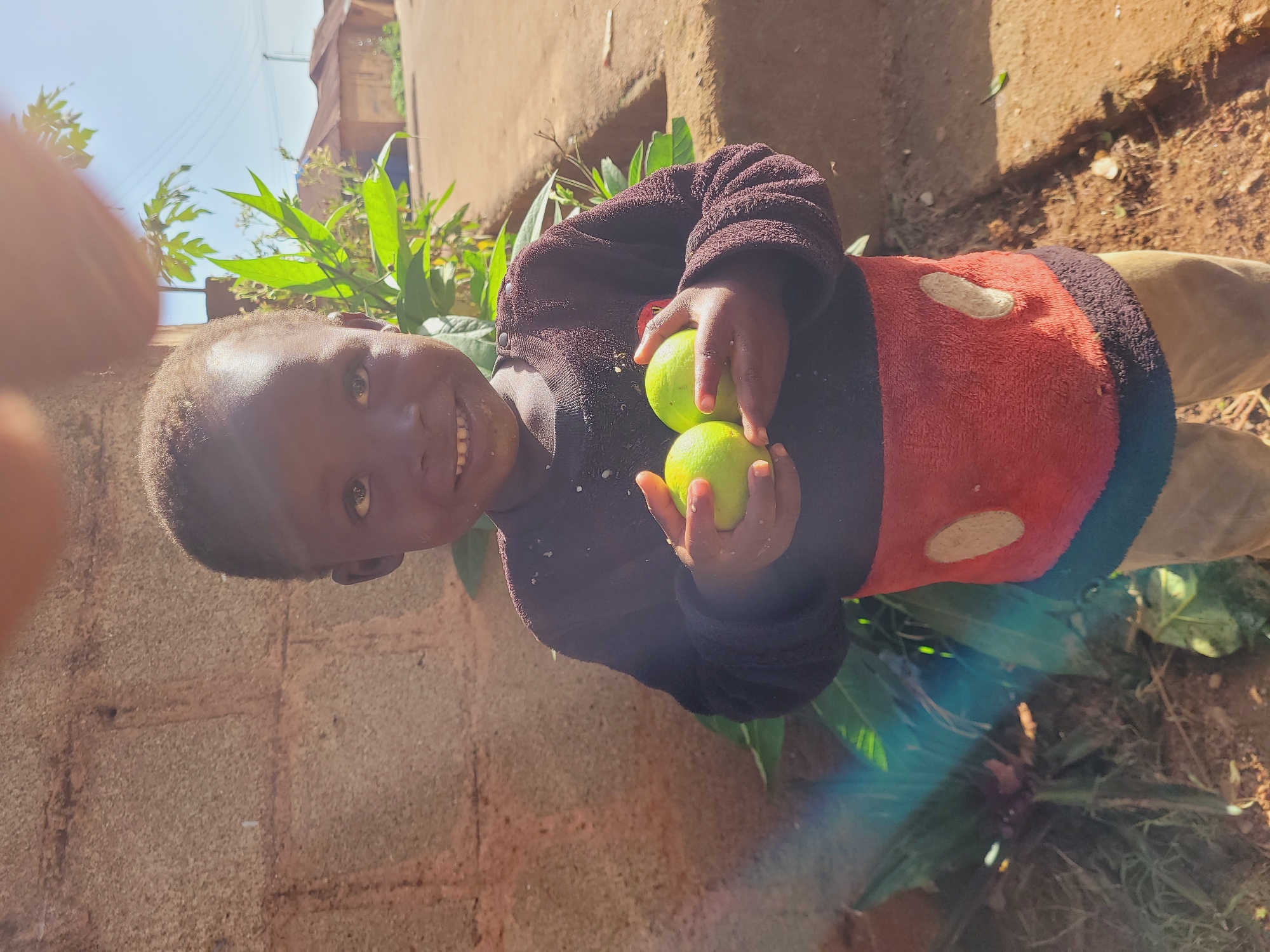 A young child with dark skin and dark hair smiles, holding two green, round fruits in their hands. The child is wearing a dark long-sleeved shirt with a red middle section featuring two oval shapes, and brown pants. The background consists of a textured brick wall to the left, lush green foliage, and a dirt path or ground with scattered leaves. In the distance, there are simple houses with brown roofs and a bright blue sky. The overall setting suggests an outdoor environment in a rural or semi-rural area, potentially in the city of Bukuru, Nigeria, given the architecture and landscape. The lighting indicates it is daytime, with sunlight creating some lens flare and highlights on the scene.