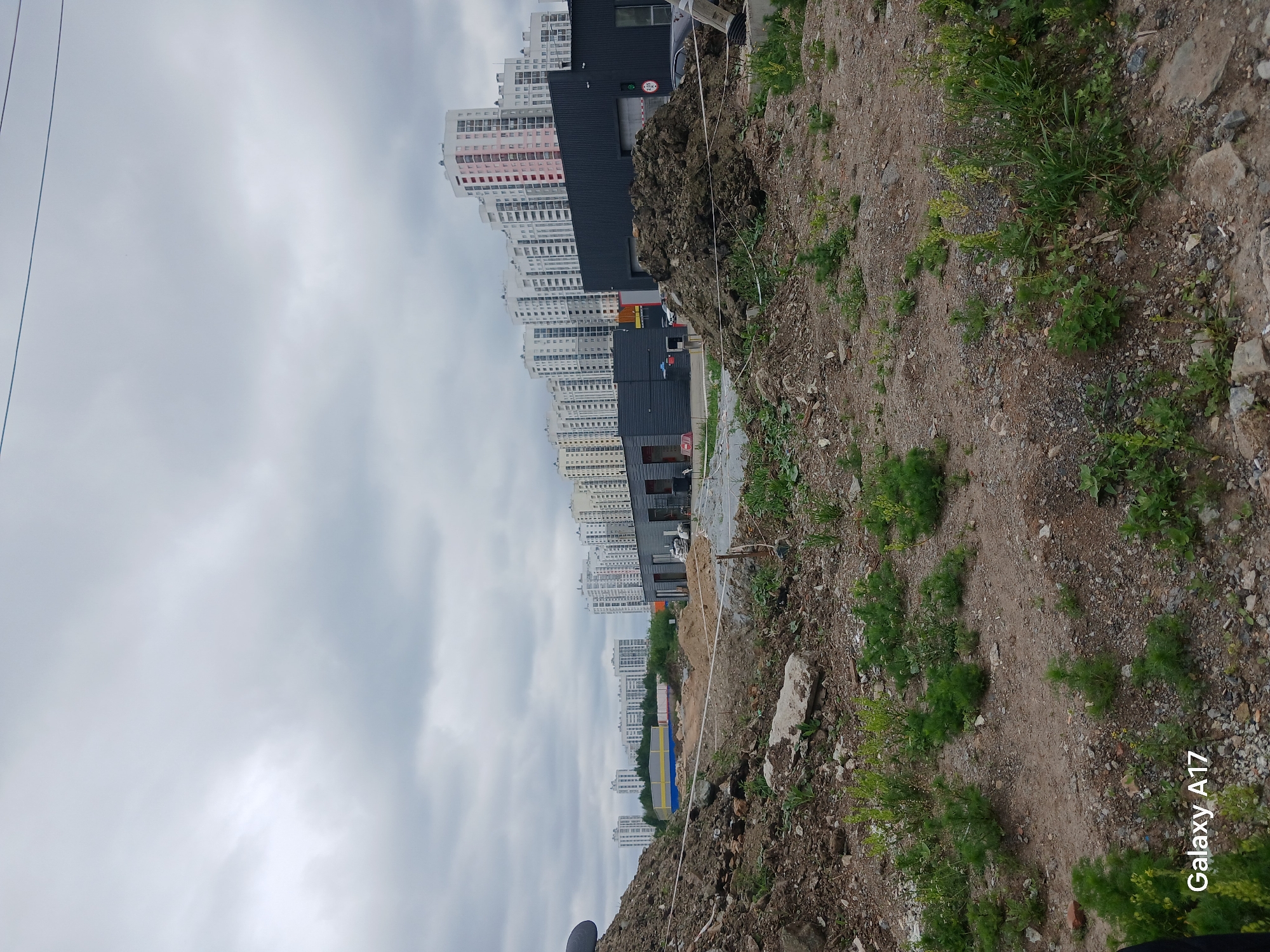 The image presents a landscape with a dirt slope in the foreground populated by sparse vegetation. In the background, a row of high-rise apartment buildings, likely located in a densely populated urban area, is visible against an overcast sky. The scene appears to be from a construction or redevelopment site. The image includes the "Galaxy A17" watermark. The location of Yekaterinburg, Russia is assumed from the urban environment and the date on the watermark. The absence of visible people and interactions indicates a static scene.