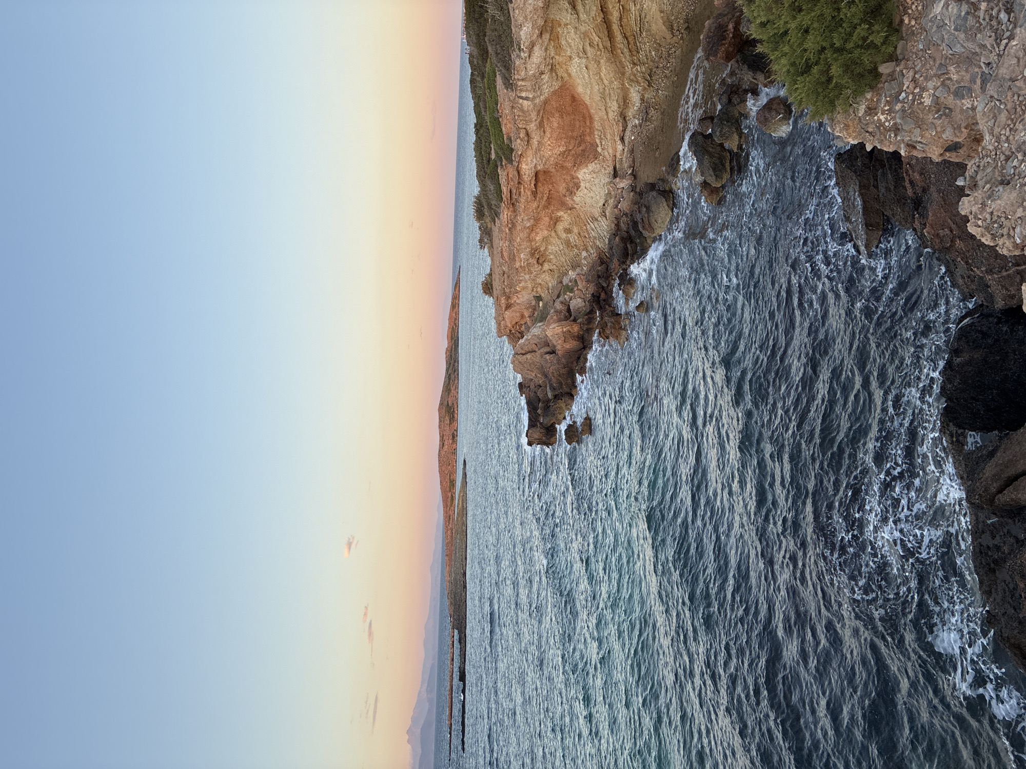 This is a vertical, eye-level shot of a rocky coastline with the sea and a sunset sky. In the foreground, dark, jagged rocks with some green shrubbery descend towards the dark blue sea, which has white foam lapping against the shore. The middle ground features a sheer cliff face of reddish-orange rock, with evidence of stratified layers and erosion. Further out, the sea stretches towards a chain of islands and a distant mountainous coastline, illuminated by the soft hues of a sunset. The sky is a gradient of pale blue at the top, transitioning to pink and orange near the horizon, with a few wispy clouds. The scene is set at the coast of Limenas Markopoulou, Greece, during the evening. There are no people or animals present.