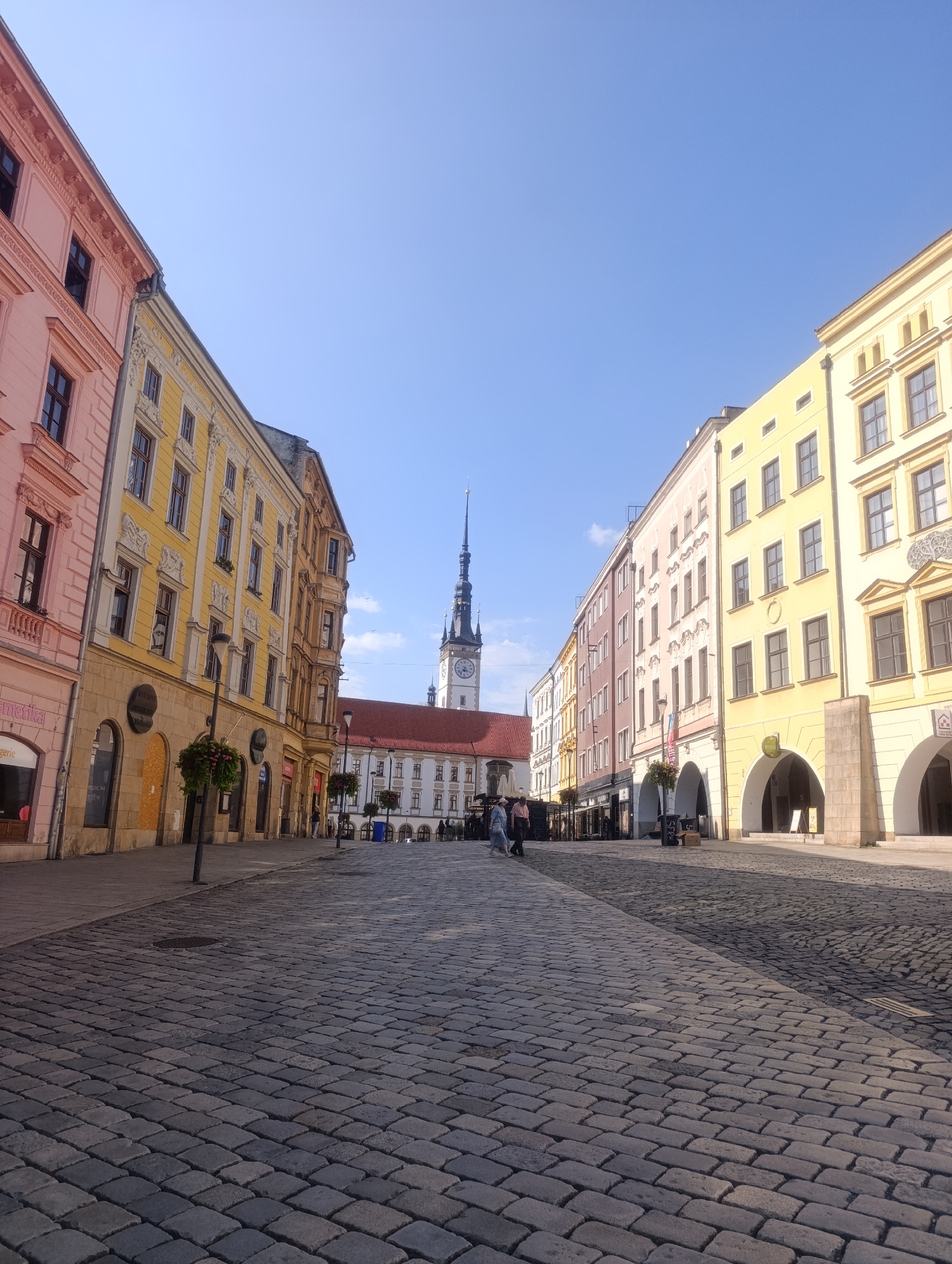 A sunlit cobblestone street in a European city is captured from a low angle, emphasizing the textured ground. Tall, colorful buildings line both sides of the street, their facades painted in shades of pink, yellow, and cream, with intricate architectural details and numerous windows. A prominent clock tower with a spire rises in the distance, centrally located within a plaza area where more buildings with red-tiled roofs are visible.
The street is relatively empty, with only a few pedestrians visible in the distance, walking casually. The sunlight casts long shadows across the cobblestones, indicating that it is likely morning or late afternoon. The sky is a clear, bright blue, suggesting fair weather. The scene evokes a sense of peacefulness and historical charm.
Some storefronts are visible at the base of the buildings on the left, with names like "Ametika" and "Lingerie" appearing in some of the shop windows. There is also some signage on the buildings with text in a foreign language, possibly Czech, given the location context of Olomouc, Czechia. The overall impression is of a picturesque and quiet urban environment bathed in pleasant sunlight.