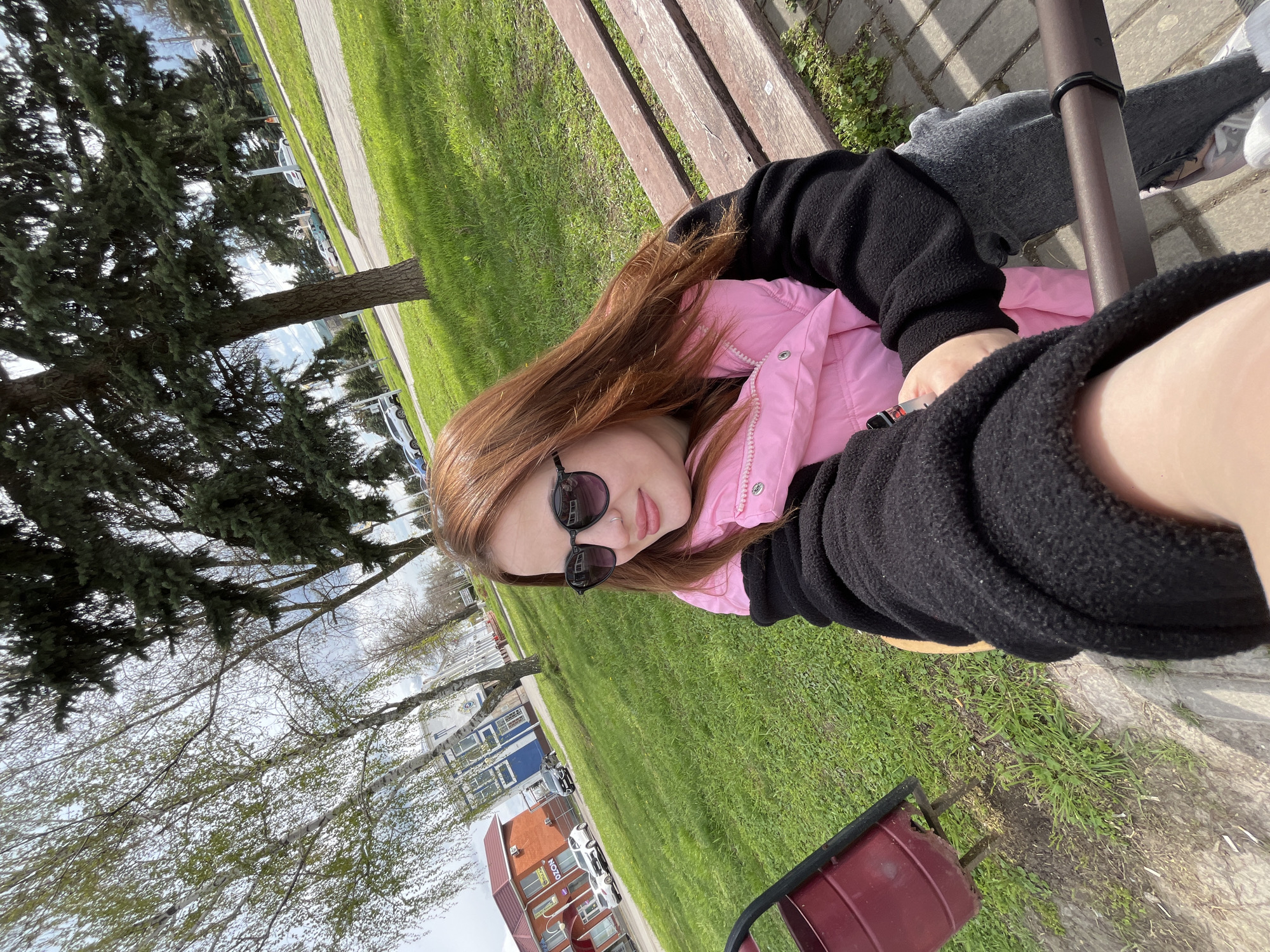 A young woman with long auburn hair and sunglasses is sitting on a wooden park bench. She is wearing a pink jacket under a black fleece. The background shows a grassy park with trees, and buildings including one with the word "NOZON" on its facade. The location appears to be Novopokrovskaya, Russia.