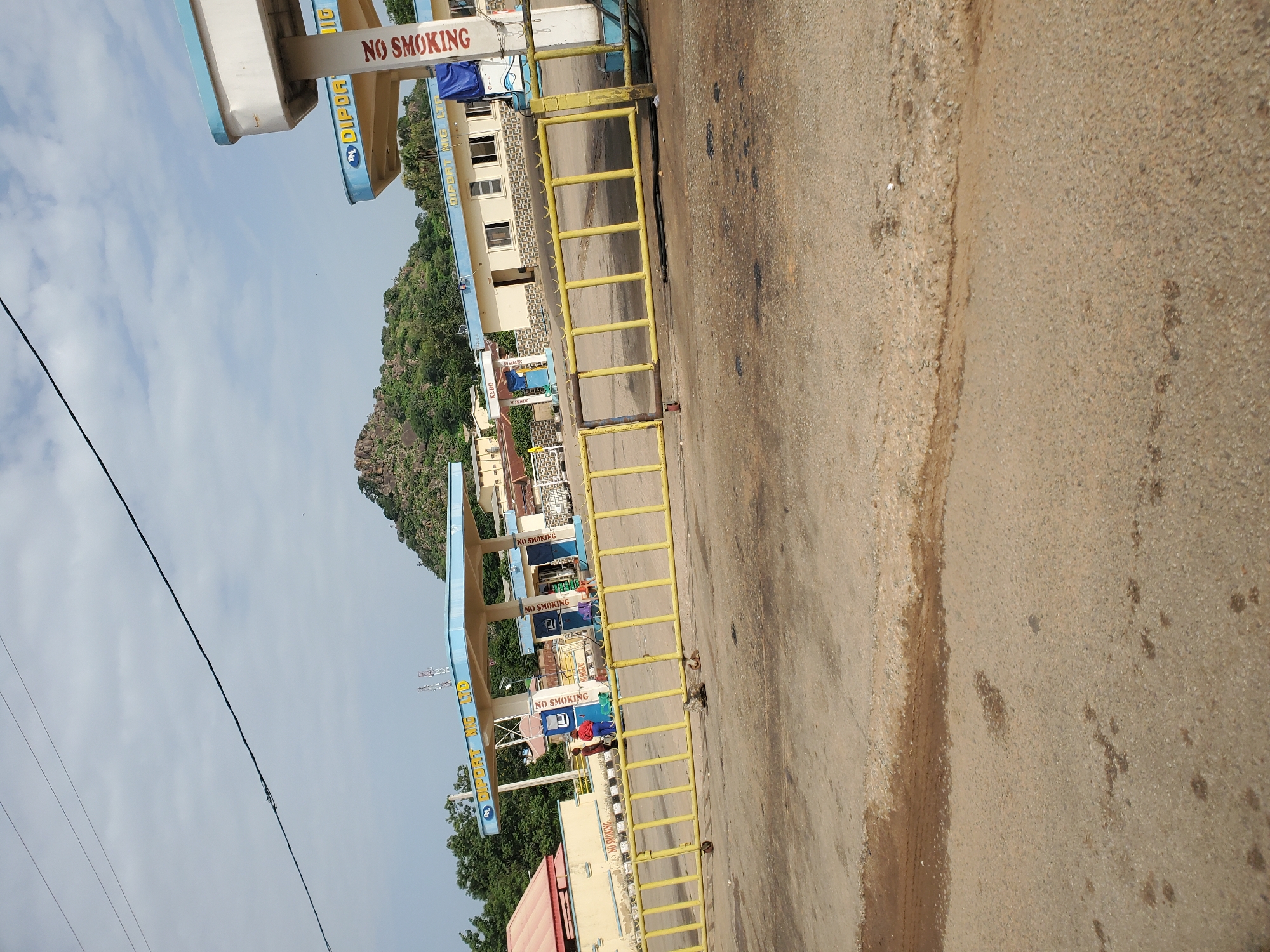 This is a medium-shot, eye-level photograph of a gas station with a yellow metal barrier in the foreground. The gas station has a blue and white canopy with signs that read "DIPORT NG LTD." and "NO SMOKING". Two people, one wearing a red shirt and the other a blue shirt, are visible near one of the gas pumps. In the background, a rocky, tree-covered hill rises under a cloudy sky. The ground in front of the gas station is paved with asphalt and appears worn and stained. This scene is located in Bukuru, Nigeria.