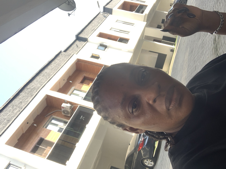 The image depicts an individual positioned in the foreground of a modern building complex located in Ikota, Nigeria. The subject, a person with dark skin and braided hair, occupies the lower half of the frame, looking directly at the camera with a neutral expression. They are wearing a dark-colored shirt. A silver-colored, chain-link bracelet is visible on their left wrist, and the fingernails of their left hand are painted black.
The background consists of at least three multi-story buildings with contemporary architecture, featuring white facades complemented by light brown, possibly wooden, paneling. Architectural details include glass balconies or railings and visible external air conditioning units. A dark-colored sedan is parked on the interlocking concrete block pavement to the left of the subject. A satellite dish is mounted on a pole against the clear, bright sky on the far right. The entire scene is brightly illuminated by natural daylight.
