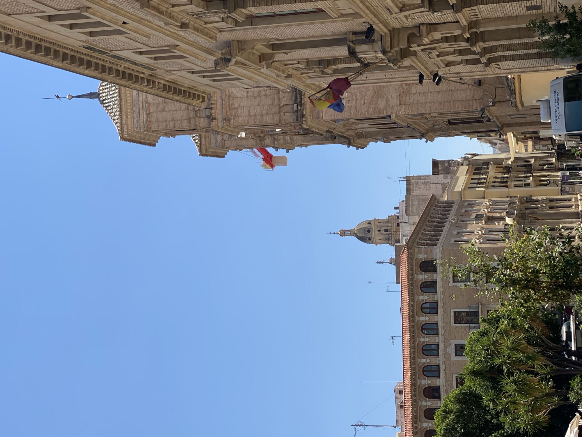 The image shows a street scene in Murcia, Spain, with a clear blue sky overhead. On the right, a tall, ornate brick building dominates the foreground, featuring classical architectural elements such as columns and arched windows. Several Spanish flags are displayed from this building. To the left, another multi-story building with arched windows and a tiled roof is visible, partially obscured by lush green trees. In the background, a distinctive domed tower rises above the cityscape, characteristic of Murcia's historical architecture. A white utility van with text indicating "OFICINA ATENCION AL CIUDADANO" (Citizen Attention Office) is parked on the street. The overall impression is of a sunny day in a historically rich urban environment.