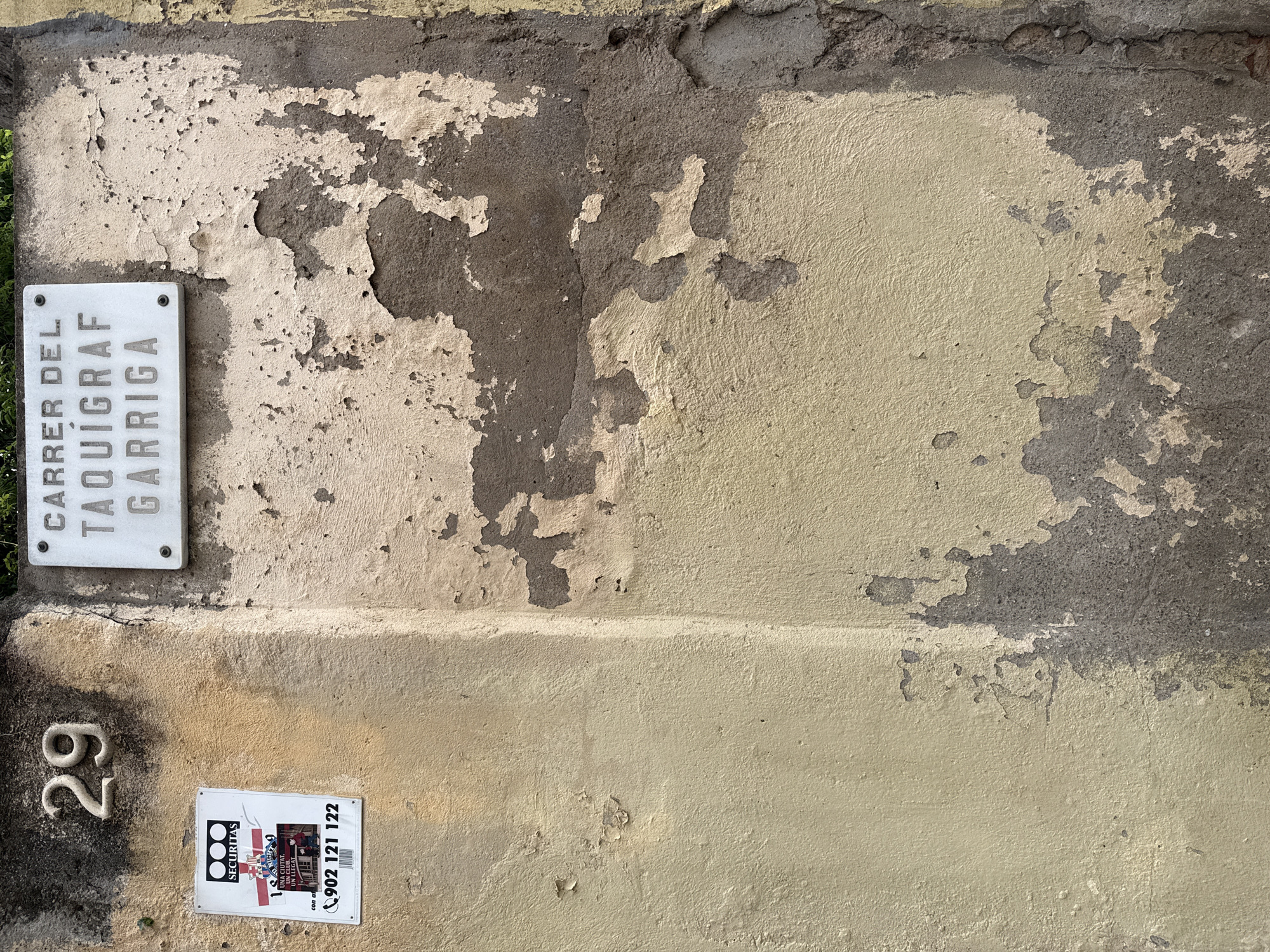 This image shows a weathered building facade in Barcelona, Spain. The wall is peeling and discolored, with patches of gray and tan. A street sign reading "Carrer del Taquígraf Garriga" is attached to the wall. Below it, a metal number "29" is mounted on the wall. Underneath the number is a "Securitas" security services advertisement sticker with the phone number 902 121 122.