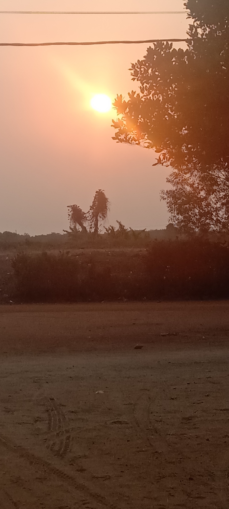 The image depicts a sunset with the sun partially obscured by trees. The sky is a warm orange hue, with visible power lines cutting across the top of the frame. In the mid-ground, there are silhouetted trees and shrubs. The foreground is a dirt or gravel surface with tire tracks visible. The overall impression is one of a rural or semi-rural landscape at dusk. There is no visible indication of the location being within Utama Jaya Mataram, Indonesia.