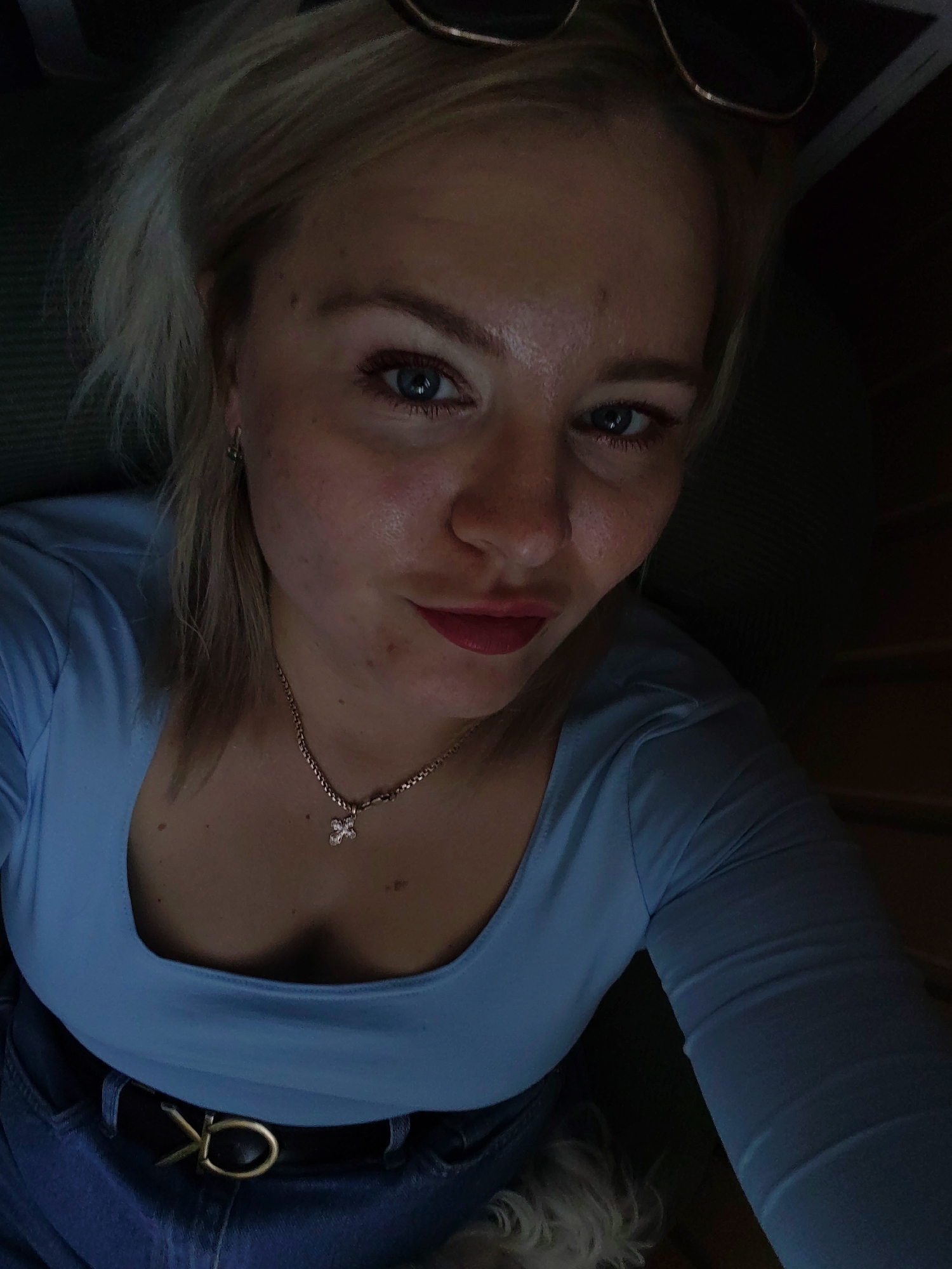 The image is a close-up, high-angle selfie of a woman with blonde hair and blue eyes, wearing a light blue long-sleeved shirt and denim jeans. She has makeup on, including lipstick, and is wearing a necklace with a cross pendant and a black belt with a gold buckle. Sunglasses are visible on top of her head. The lighting suggests an indoor setting with a dark background. There is no visible indication of the city of Izhevsk, Russia.