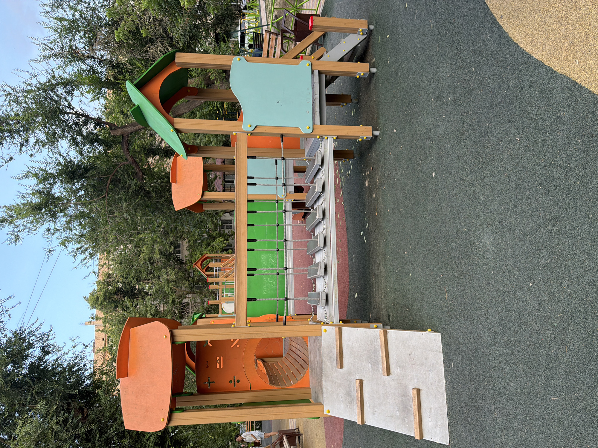 The image depicts a vibrant outdoor children's playground in Shymkent, Kazakhstan. The main subject is a multi-level wooden playground structure with bright orange, green, and light blue accents. It features various elements such as slides, climbing ropes, and platforms, designed for children's recreation. The playground is situated on a safe, shock-absorbent rubberized surface, with sections in dark green and a lighter beige, indicating different zones or age-appropriateness. Trees with lush green foliage surround the playground, suggesting a pleasant outdoor environment. The sky visible in the upper left corner is a clear blue, hinting at fair weather. In the background, a building with light-colored bricks can be seen through the trees, providing context of an urban or suburban setting. A portion of a light brown wooden bench is visible in the lower left corner of the frame.
While no specific activity is captured in action, the playground's design implies it is a place for children to play and engage in physical activities like climbing, sliding, and imaginative play. The overall atmosphere appears to be peaceful and well-maintained. The time of day is likely daytime, with the sun casting shadows, particularly a large shadow from the playground structure on the right side of the frame. No people are clearly visible engaged in play within the primary focus of the image, though a blurred figure can be seen in the lower left. No visible text is present on the playground equipment or surrounding areas.