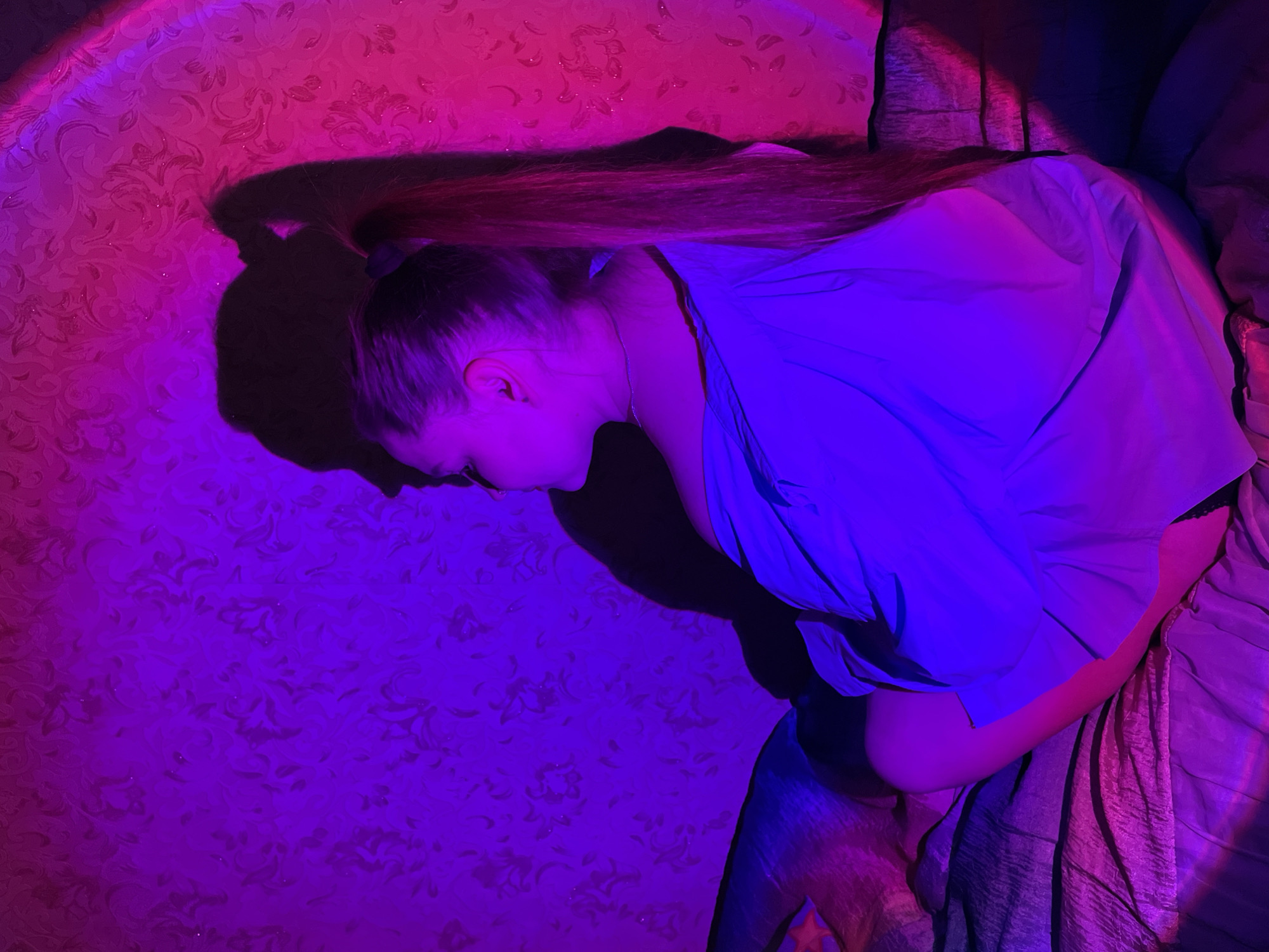 An overhead shot captures a young woman lying horizontally, bathed in dramatic magenta and blue-purple lighting. Her head is positioned towards the left of the frame, with her face turned downwards, partially obscured, suggesting rest or introspection. She has long, dark hair tied in a ponytail, which extends significantly behind her head. A prominent, sharp shadow of her head and ponytail is cast on the patterned wall to her left.
She is wearing a loose, light-colored top, possibly a shirt, and light-colored bottoms, both appearing indigo/violet due to the strong blue-purple light. The magenta light illuminates the wall behind her head, revealing an intricate floral or damask pattern. A subtle silver chain is visible around her neck. She is lying on a textured, crumpled fabric, likely a bed or sofa covering, which also reflects the colored lights.
The scene appears to be indoors, likely at night or in a room with controlled artificial lighting. The combination of intense magenta and blue-purple lights creates a vibrant, almost ethereal atmosphere, with a strong color gradient across the image. No visible text is present in the image. The setting is in Izhevsk, Russia.