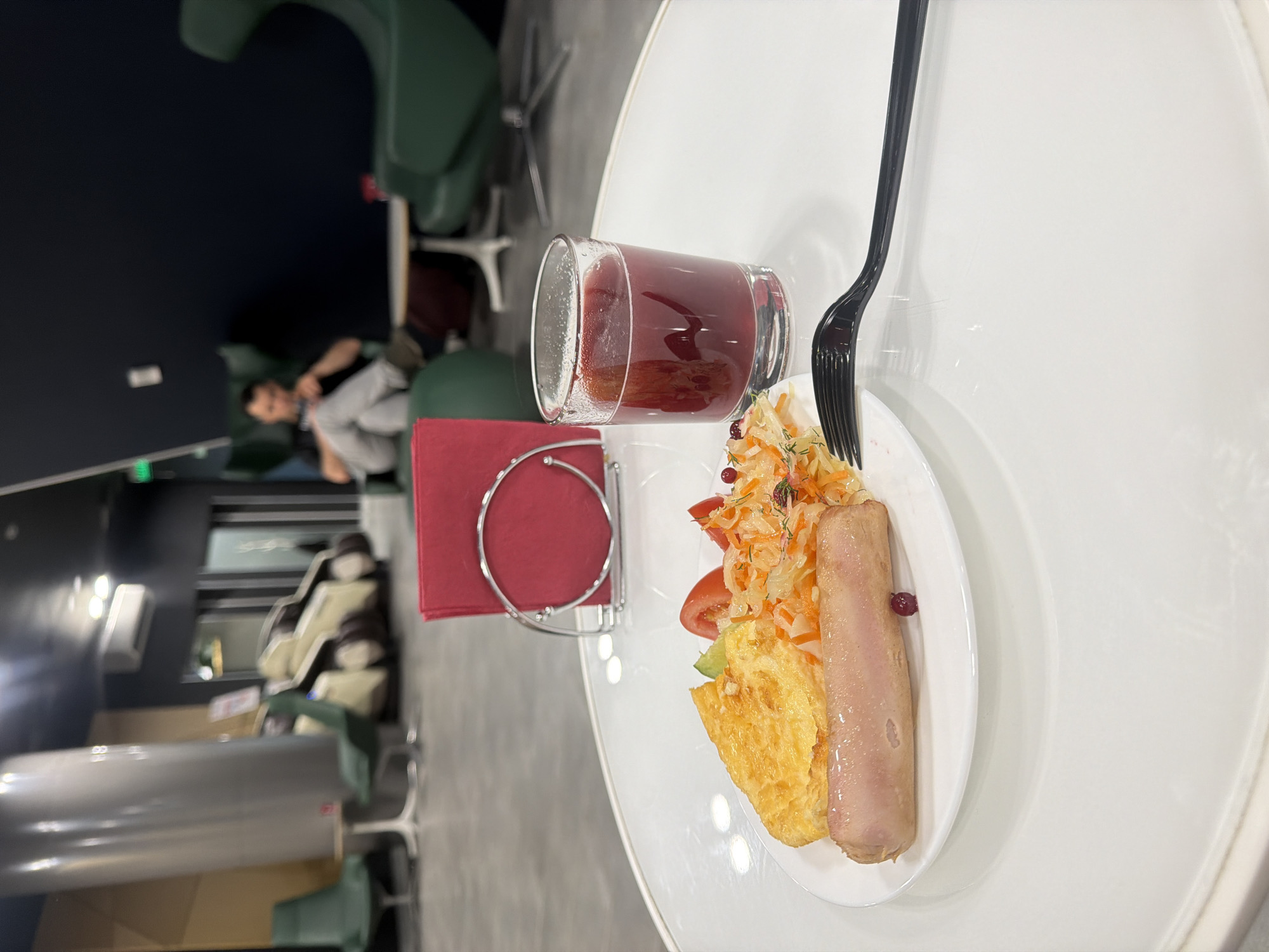 The image depicts a plate of food and a glass of red liquid on a white table. The food appears to consist of sausage, scrambled eggs, and a salad with tomatoes and sauerkraut. A red napkin in a silver holder sits to the left of the plate. A black fork is placed on the plate.

In the background, there is a waiting area or lounge with various types of seating, including chairs with high backs and curved seating. A person is visible in the background, seated in one of the chairs. 

The image location is the city of Kultaevo, Russia.