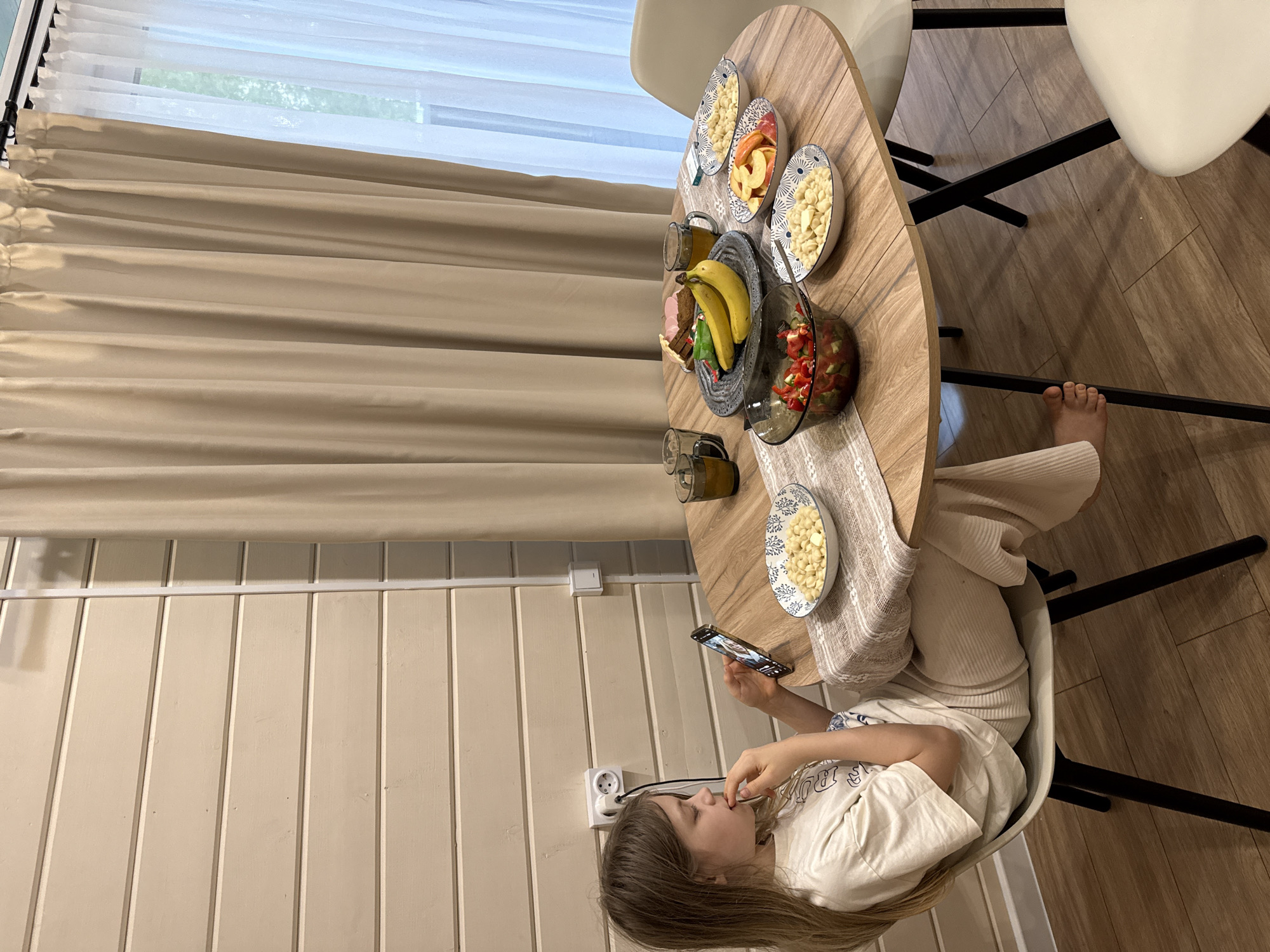 A girl is sitting at a round wooden table with a white table runner and a variety of food items: bananas, a bowl of salad, fruit slices, and small portions of what appears to be pasta in bowls. She is using a smartphone in her hand while eating a small portion from the table. She's wearing beige colored pants and a white T-shirt. The table is surrounded by white chairs with black legs. The room has wooden walls and a large window covered with beige curtains and sheer white curtains. The floor is wooden. The picture appears to have been taken indoors during daytime in Buzhaninovo, Russia.