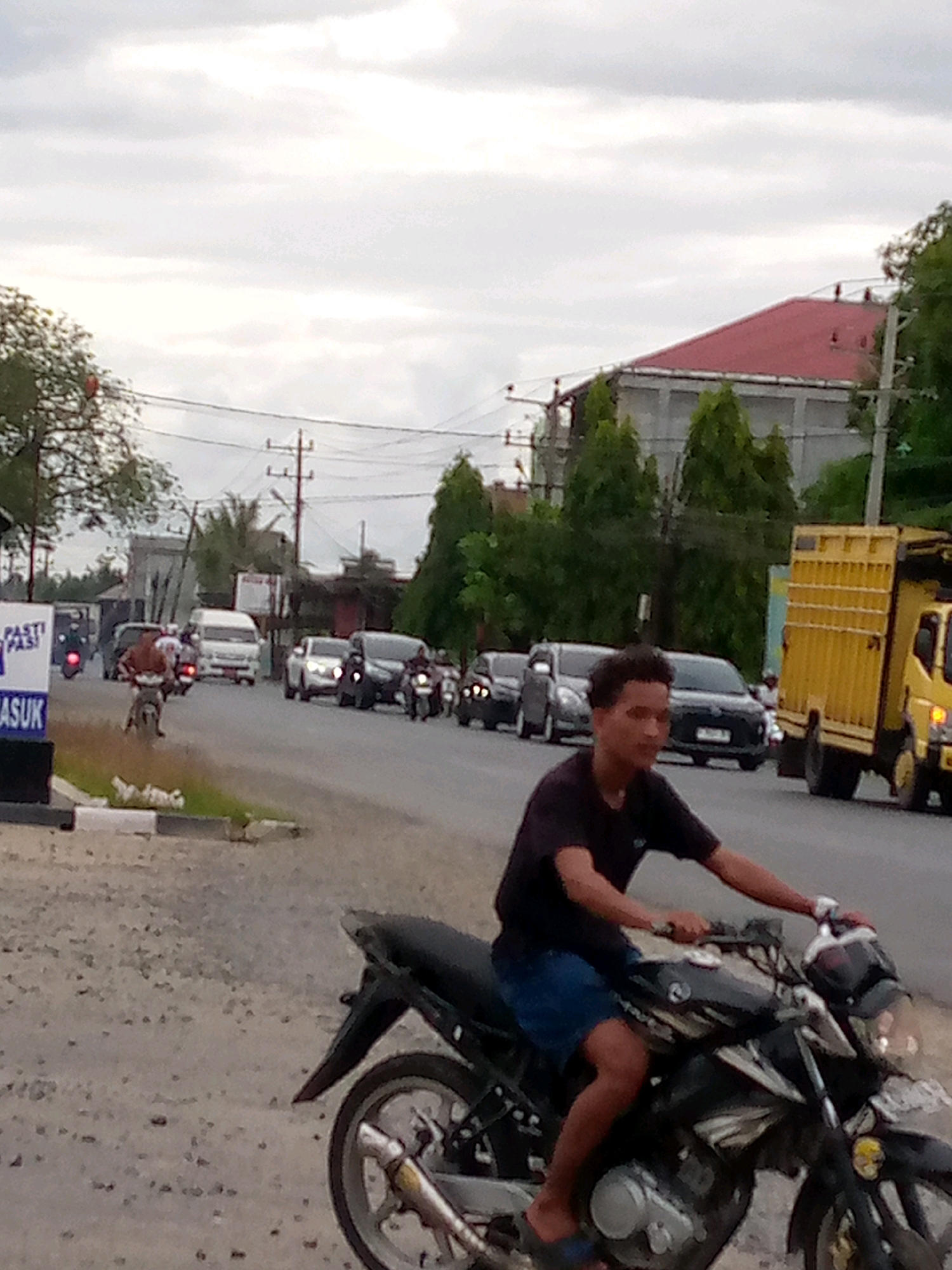 A young man rides a black motorcycle on a dirt and gravel road, in what appears to be a semi-urban or rural setting in Keudebayu, Indonesia. He is wearing a black t-shirt and blue shorts, with his right arm extended and hand on the handlebars. The motorcycle's front wheel and part of the engine are visible. In the background, a busy street with several cars and motorcycles is seen, along with trees, buildings, and utility poles. A yellow truck with a covered cargo bed is parked on the right side of the road. The sky is overcast with clouds.