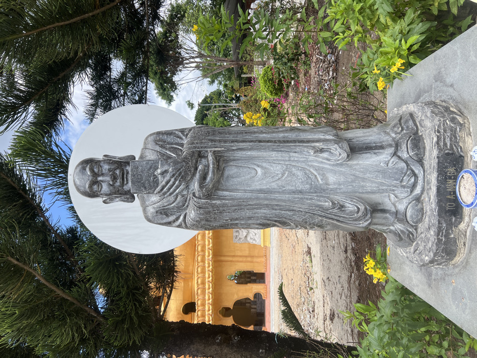 A tall, grey stone statue of a Buddhist monk, identified as "TÔ A-NAN-ĐA" on its pedestal, stands centrally in an outdoor setting within the city of Đăk Mar, Vietnam. The monk statue has a serene expression, elongated earlobes, and holds a rectangular object with both hands in front of its chest. A large, white circular disc serves as a backdrop behind the monk's head. To the statue's left, a multi-tiered, golden-yellow temple building with ornate architectural details and arched openings is visible; a dark silhouette of a seated Buddha statue is discernible within one archway. The ground in front of the temple building is unpaved earth. To the statue's right and in the mid-background, a landscaped garden features various green shrubs, trees, and flowering plants with yellow and pink blossoms. The upper quadrants of the image are partially framed by green pine tree branches. The sky above is partly cloudy blue. At the base of the statue's pedestal, a small blue and white ceramic bowl containing ash or sand, along with two smaller white objects, is present.