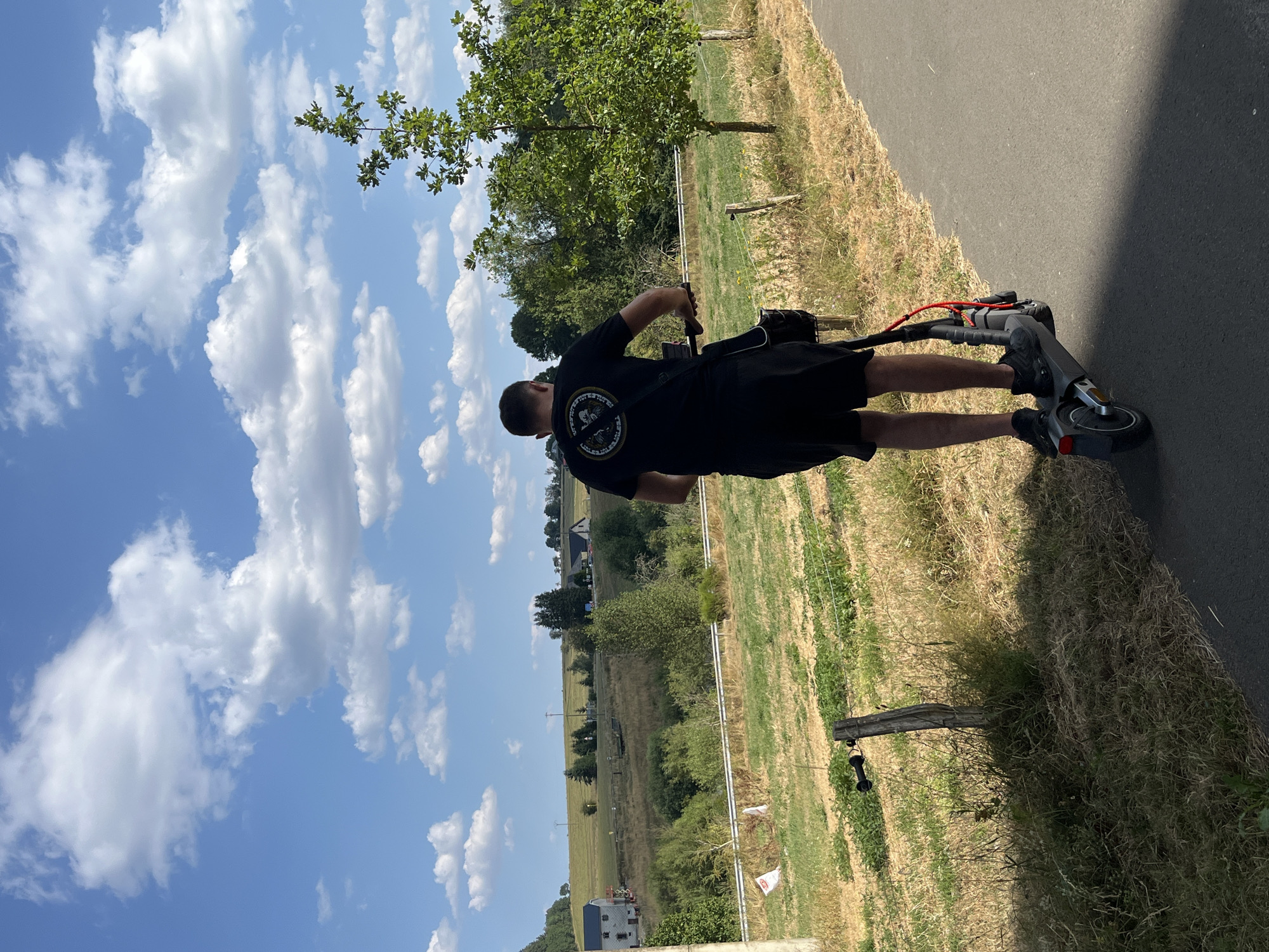 A man is riding an electric scooter on a paved path in Saint-Vith, Belgium. He is wearing a black t-shirt and black shorts, and a black bag is slung over his shoulder. The path is bordered by dry grass and a wooden fence. In the background, there is a green, hilly landscape with scattered trees and houses. The sky is blue with white, fluffy clouds.