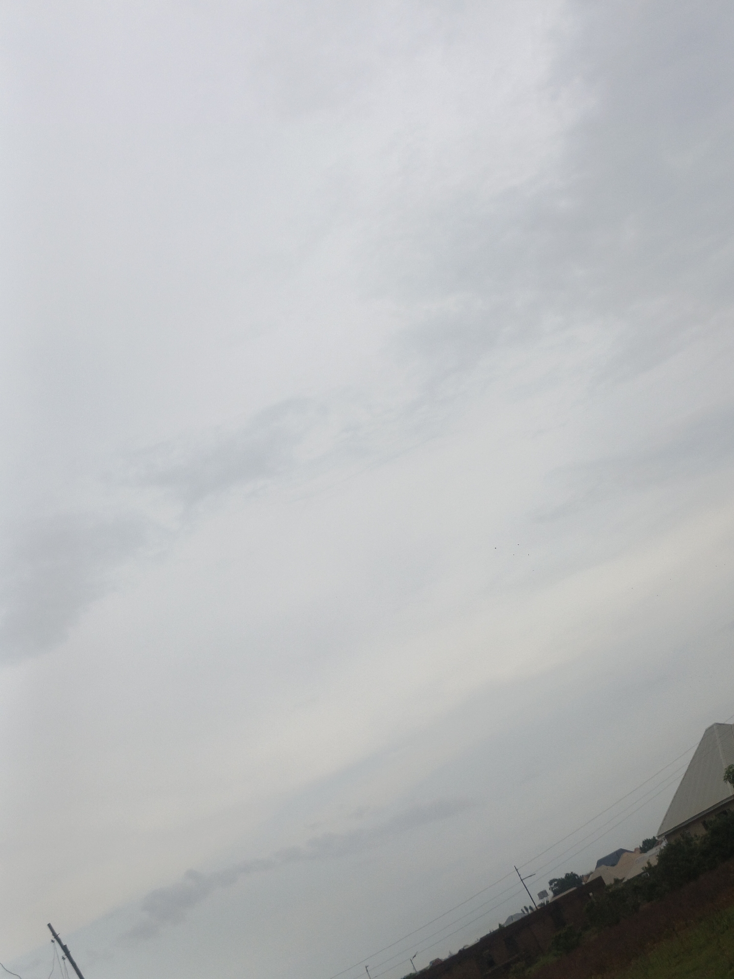 The image captures an overcast sky with scattered clouds, suggesting a diffused light or pre-rain conditions. A power pole with wires is visible in the lower left quadrant, angled upwards. The middle ground shows a line of buildings and vegetation. To the right, a large, light-colored, angular structure dominates the skyline, possibly a modern warehouse or industrial building. The ground appears to be a mix of reddish-brown soil and green grass. The scene is situated in Rafin Bauna, Nigeria, indicating a semi-urban or peri-urban landscape. There are no people or discernible activities in the image.
