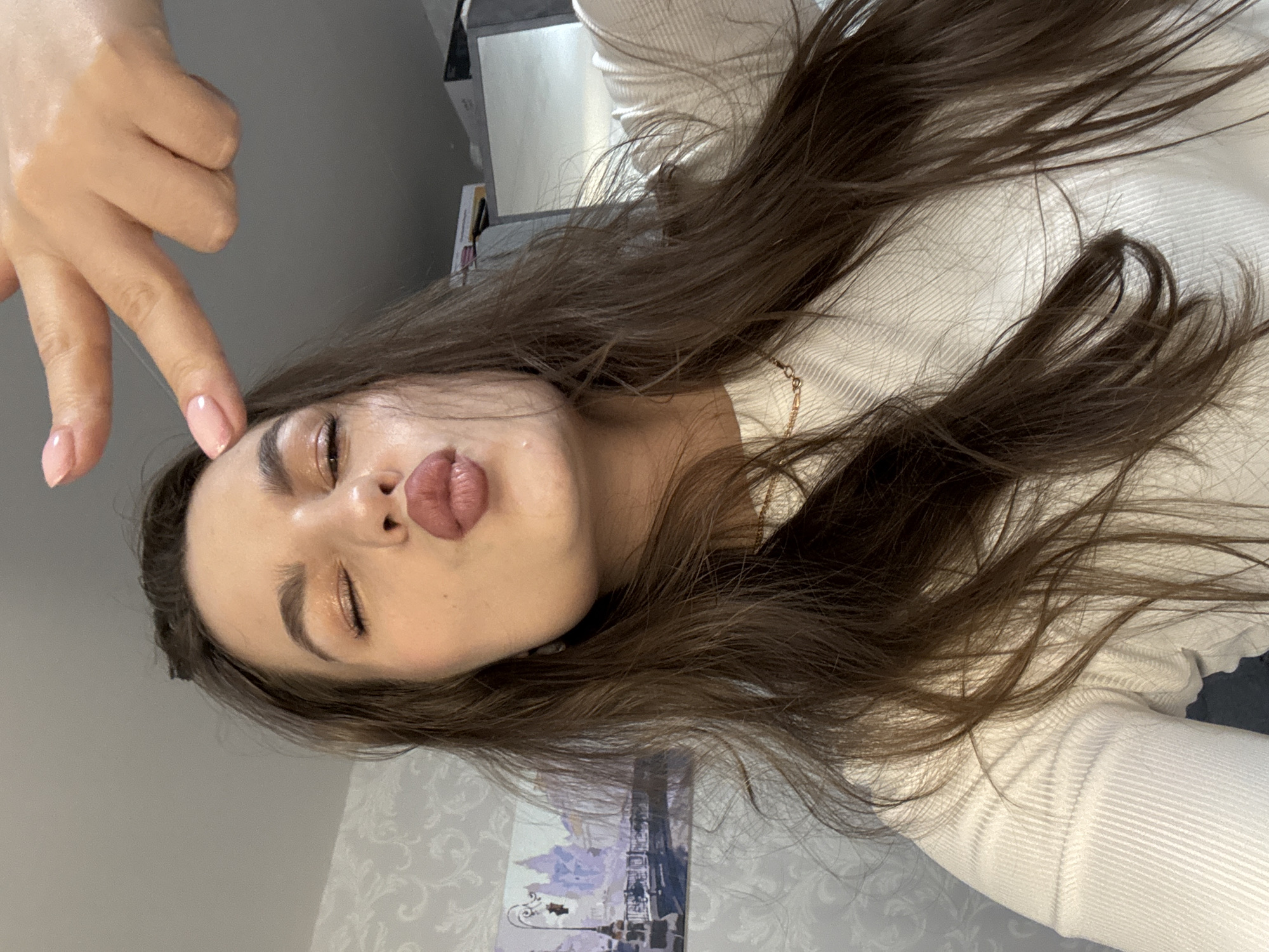 The image is a close-up selfie of a young woman taken in an indoor setting in Chaikovsky, Russia. The woman, with long, wavy dark brown hair fanned around her, is looking towards the camera with her head tilted. She has her eyes closed or heavily lidded and her lips puckered in a playful "kissing" or "duck face" expression. Her left hand is visible in the upper left quadrant, with her index and middle fingers extended and lightly touching her temple area.
She is wearing a light-colored, likely white or off-white, ribbed long-sleeved top or sweater. A delicate gold chain necklace is visible around her neck. Her nails are manicured with a light pink or nude polish. She has visible makeup, including shimmering eyeshadow and lip gloss, enhancing her features.
The background shows parts of a room. A plain light grey wall is visible on the left, and a wall with a subtle, light-toned patterned wallpaper is seen on the right. Behind her hair, a small framed artwork depicting an urban landscape with buildings and a lamppost is visible. No text is visible in the image. The overall impression is a casual, lighthearted self-portrait.