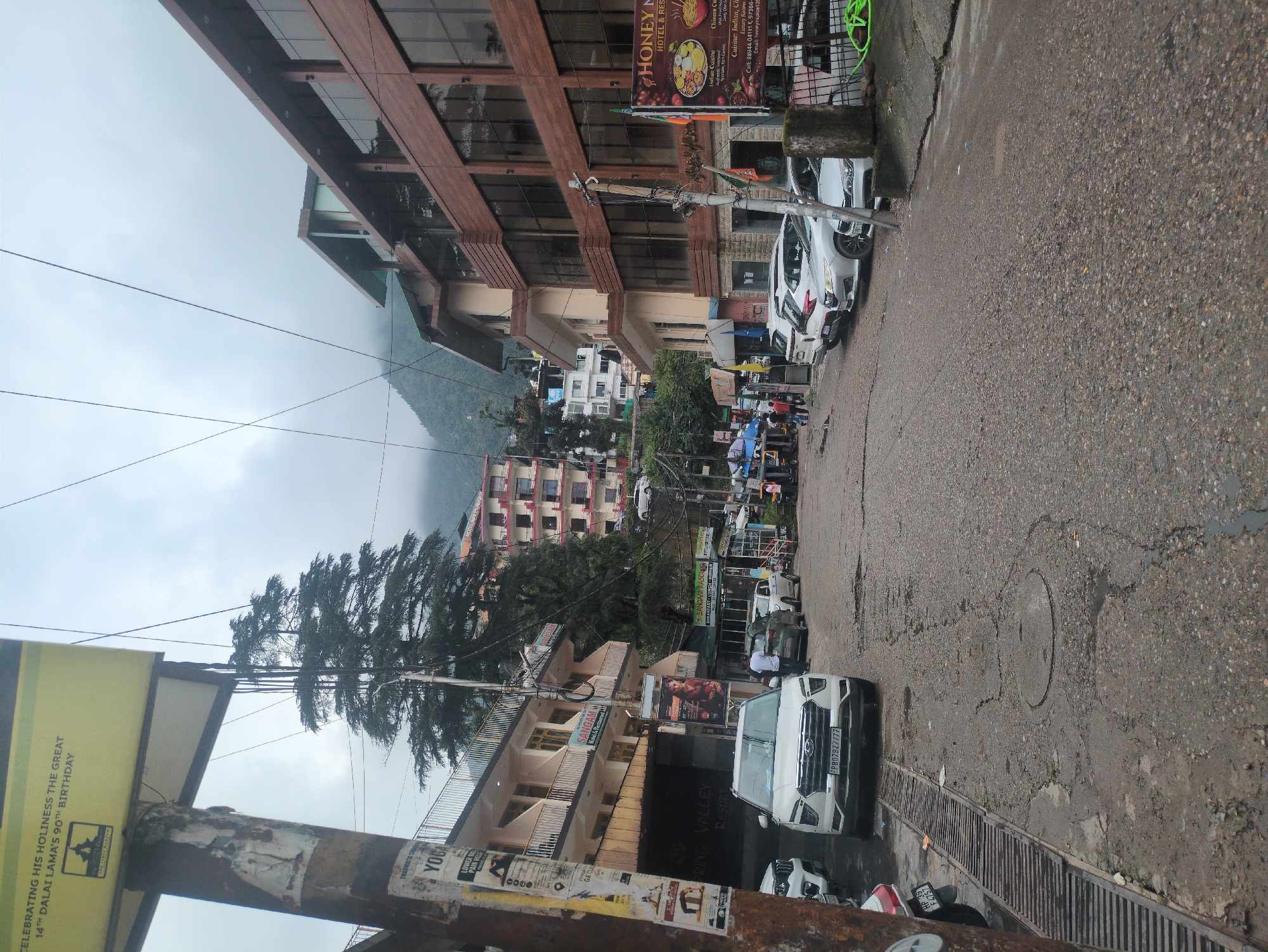 The image depicts a street scene in Bhagsu, India, under an overcast sky. The street is paved with asphalt and shows signs of wear, with visible potholes and a metal grate along the left side. Several white cars are parked along the right side of the street.
On the left side of the street, a multi-story building with balconies is visible, featuring signs for "SANGAM HOTEL & RESTAURANT". A large yellow billboard is mounted on a pole in the foreground, advertising "CELEBRATING HIS HOLINESS THE GREAT 14th DALAI LAMA'S 90th BIRTHDAY". Beneath this, another pole has various smaller posters, one of which reads "YOGA".
On the right side, a modern building with a brown wooden facade and large windows dominates the scene. A banner for "HONEY ROCKS HOTEL & RESTAURANT" is displayed on this building, featuring images of food. Further down the street, more buildings are visible, including a multi-colored residential or hotel building.
A few individuals are present on the street, some walking and others standing near the parked cars. A rickshaw is visible in the distance on the left. The overall impression is of a busy, commercial street in a hilly town.