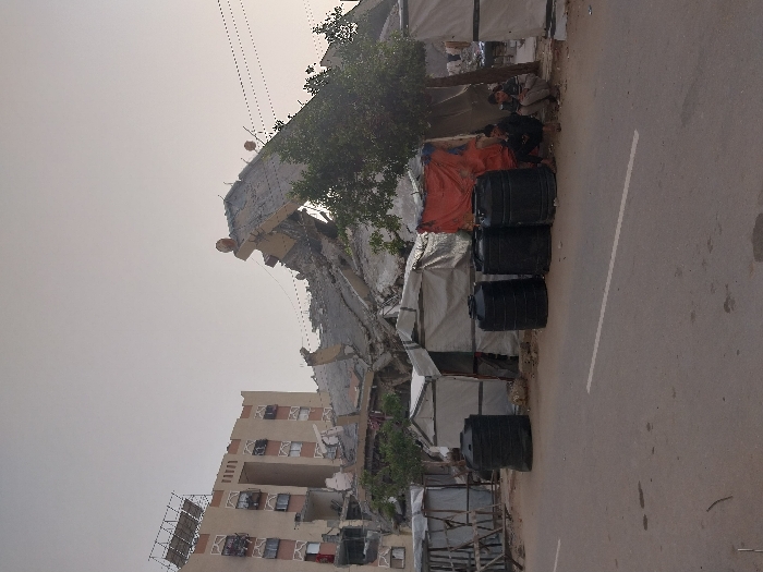 The image depicts a street scene in Khan Yunis, Palestinian Territory, characterized by signs of displacement and structural damage. In the foreground, an asphalt road with a single solid white line runs horizontally across the lower third of the frame. Immediately adjacent to the road, on uneven dirt ground, a row of five black plastic barrels is visible. Behind these barrels, a series of makeshift shelters constructed from tarpaulins (white, grey, and orange) and wooden frames are erected. Two individuals are partially visible to the left of the barrels, near these shelters; one wears dark clothing, the other lighter attire. To the left-midground, a large pile of concrete rubble and twisted rebar indicates a collapsed or heavily damaged building, with a small green tree growing amidst the debris. Electrical wires with circular insulators traverse the mid-distance. In the background on the right, a multi-story residential building, light brown in color, stands largely intact, featuring numerous windows and balconies. A metal framework structure is visible on its roofline. The sky above is pale and appears hazy or overcast.