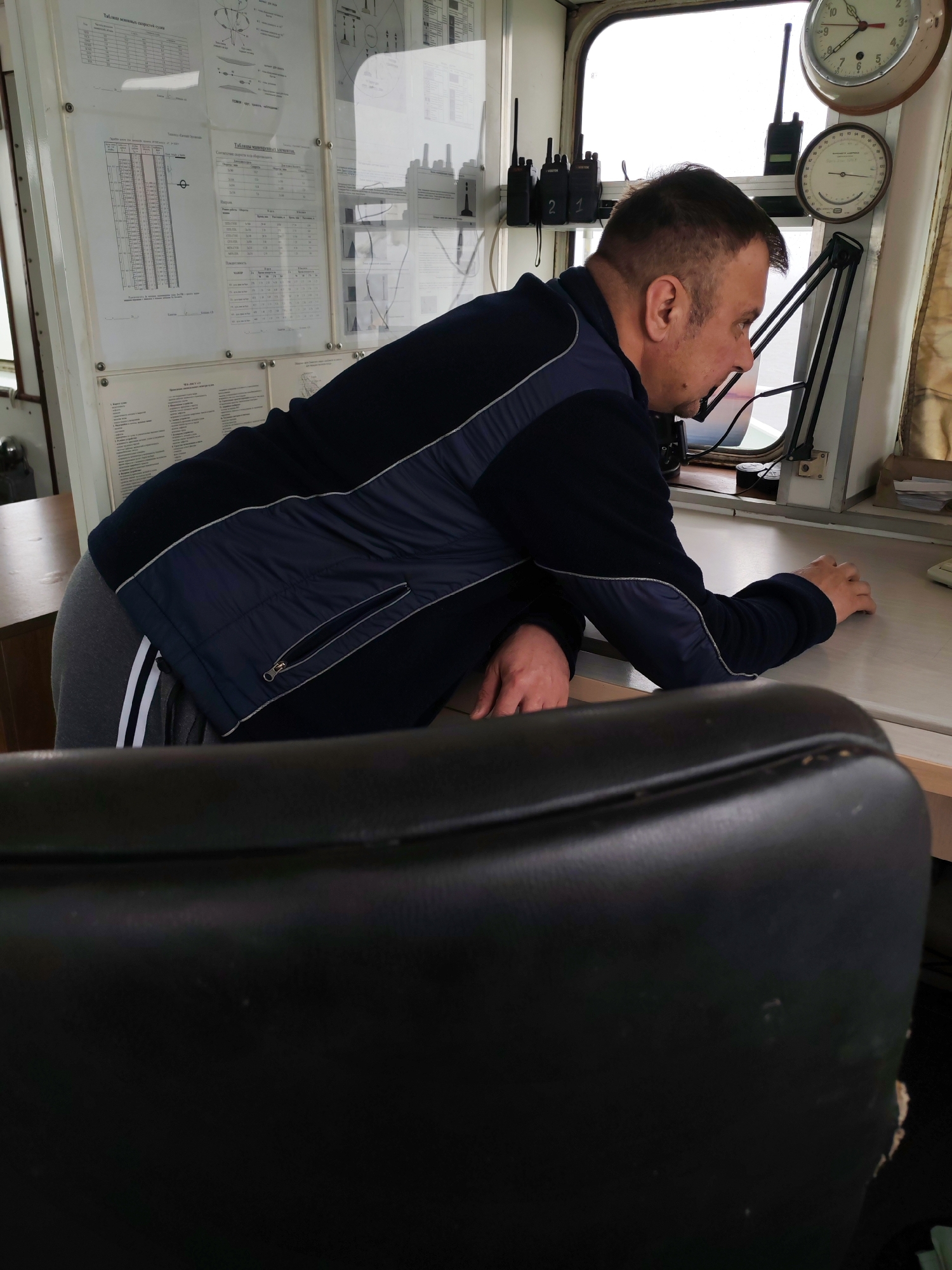This photograph captures a man intently focused on an activity within what appears to be the bridge or control room of a vessel, located in Shcheleiki, Russia. The man, seen in profile, is middle-aged with short dark hair, wearing a dark blue and grey jacket over grey track pants with white stripes. He is leaning forward over a light-colored desk or console, with his left forearm resting on it and his right hand partially visible.
The scene is functional and practical. Behind the man, a white panel displays several documents under clear protective sheets. These documents include technical charts and tables, notably titled in Cyrillic, such as "Таблица возможных скоростей судна" (Table of possible vessel speeds), "Проба воды для измерения глубины и груз в" (Water sample for measuring depth and cargo in), and "Таблицы маневренных элементов" (Tables of maneuvering elements).
On a shelf above the documents, three walkie-talkies are visible, one marked "2 1." Above the console, a round clock with Roman numerals shows approximately 12:45, and a smaller round gauge, possibly a barometer, is mounted below it. A black adjustable arm, likely for a lamp or monitor, extends from the desk. A large window behind the man reveals an overcast, bright sky, indicating daytime with dull weather. The man appears focused and engaged in his task, possibly monitoring navigation or operations. In the immediate foreground, the back of a worn black leather-like office chair is visible, partially obscuring the desk.